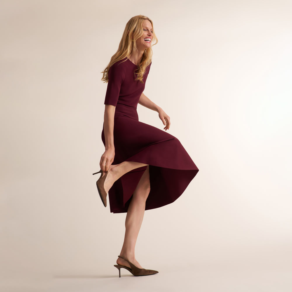 A woman modeling a fitted, knee-length burgundy dress with elbow-length sleeves and a flared skirt, posed mid-step against a plain warm beige studio background.