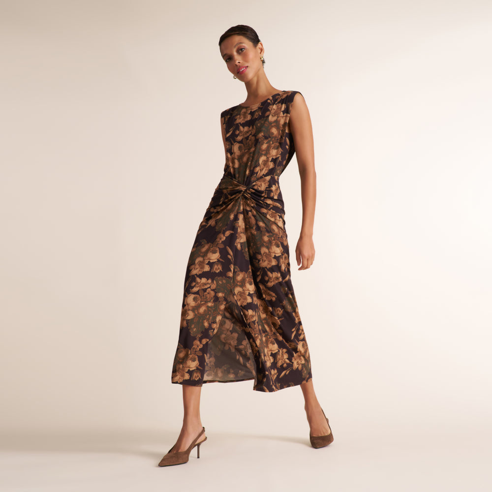 A woman models a sleeveless midi dress with a dark brown and black floral pattern and a gathered waist detail, paired with brown pointed-toe heels against a plain beige studio background.
