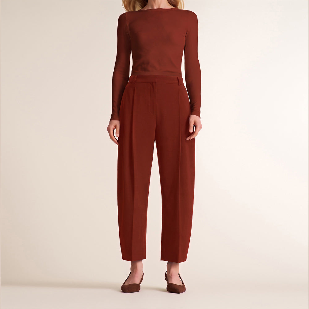 A woman modeling a rust-brown two-piece set consisting of a long-sleeve top and matching high-waisted cropped trousers, paired with brown flats, against a plain beige studio background.