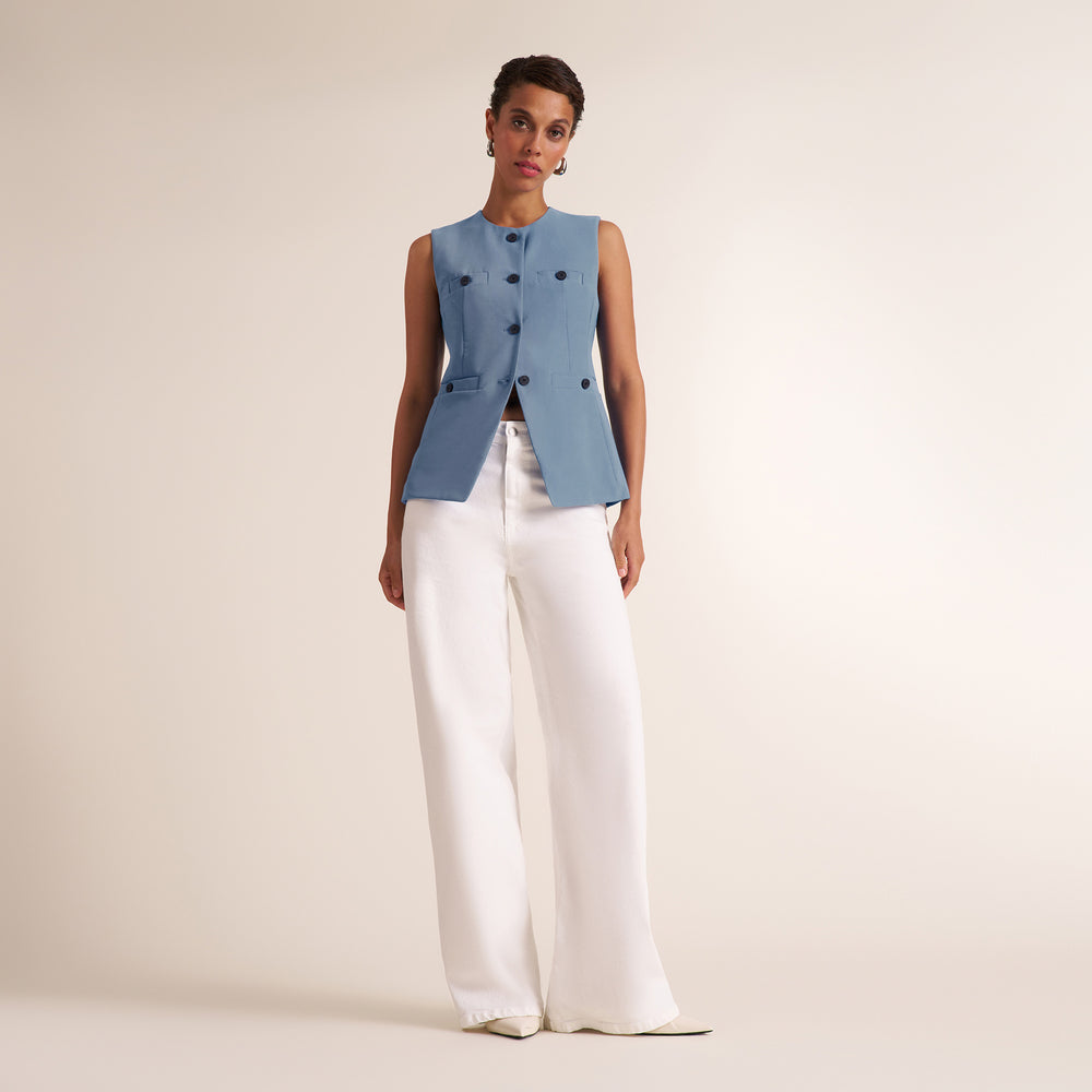 A model stands against a warm beige studio background wearing a sleeveless light blue vest with dark buttons over a black top and white wide-leg trousers.