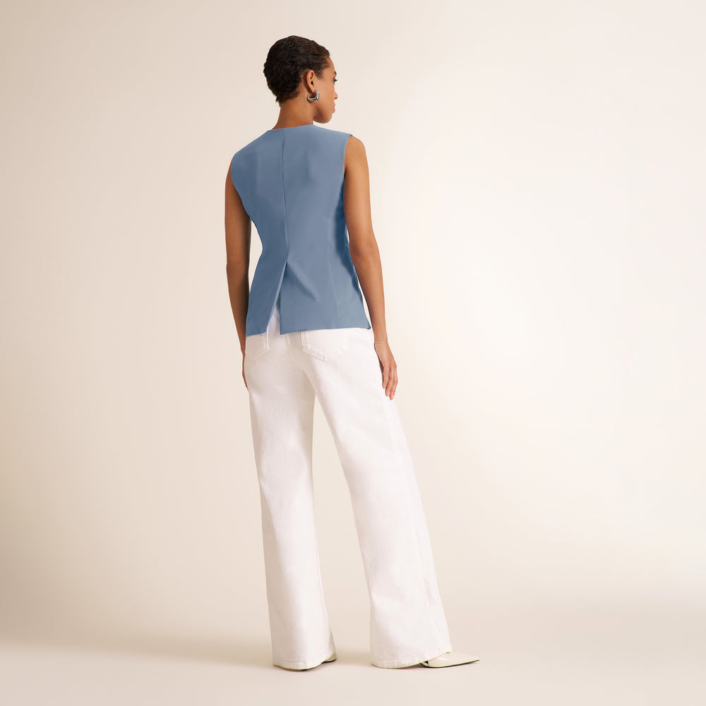A woman shown from the back wearing a sleeveless light blue top and white wide-leg pants, standing against a plain warm off-white studio background.