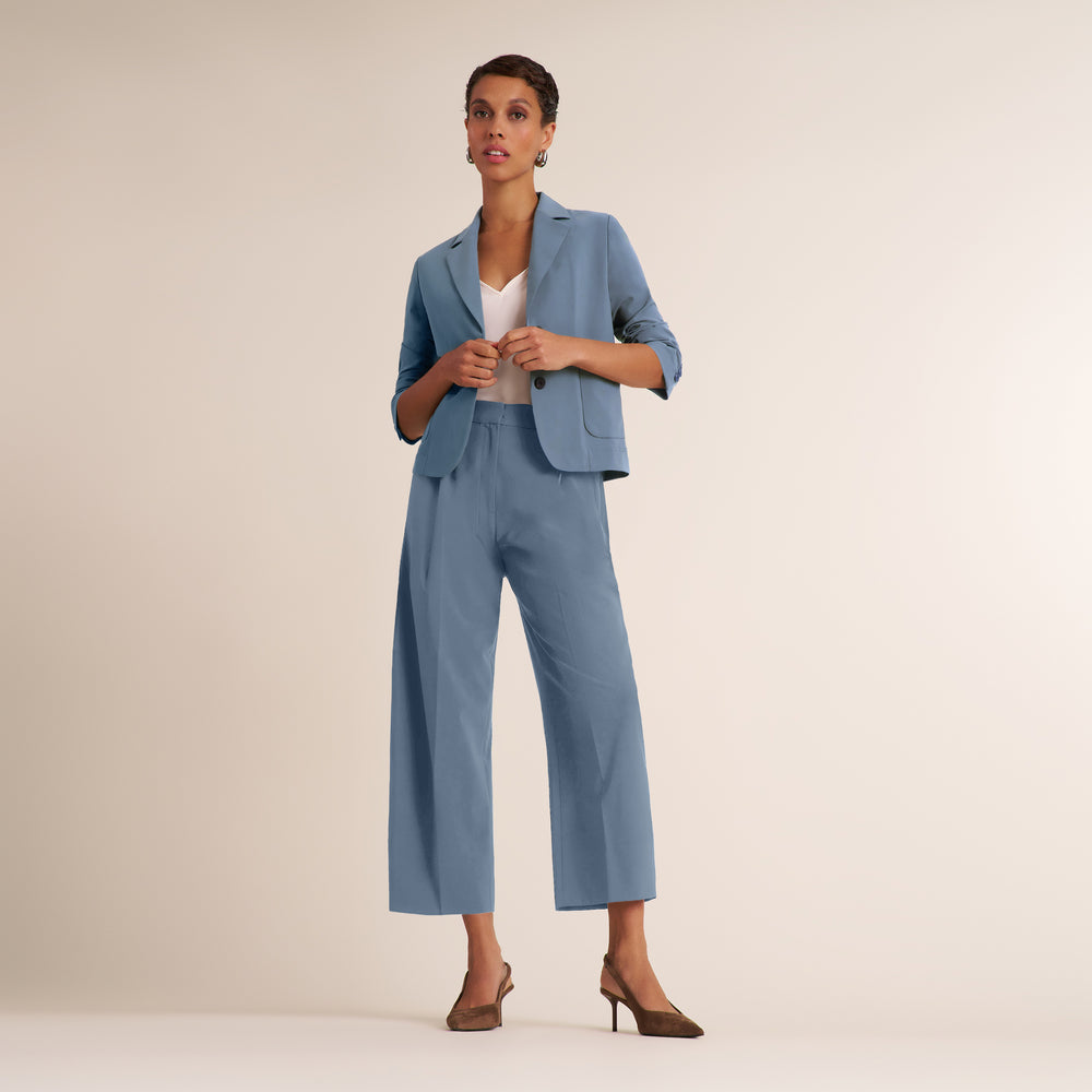 A woman modeling a light blue cropped blazer and matching wide-leg cropped trousers with a white top and brown heels, standing against a plain beige studio background.