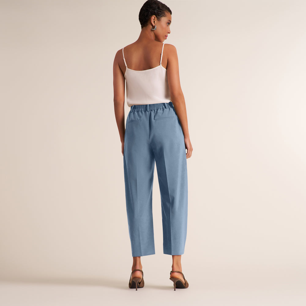 A woman is shown from the back wearing light blue cropped wide-leg pants, a white spaghetti-strap camisole, and brown heeled sandals against a plain beige studio background.
