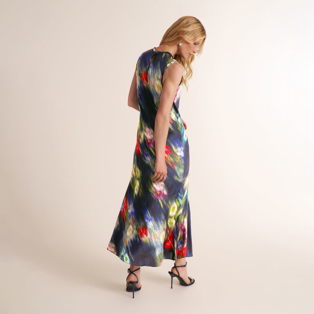 A woman modeling a sleeveless maxi dress with a dark navy base and a vibrant, multicolored abstract floral print, shown from a three-quarters back view against a plain beige studio background.