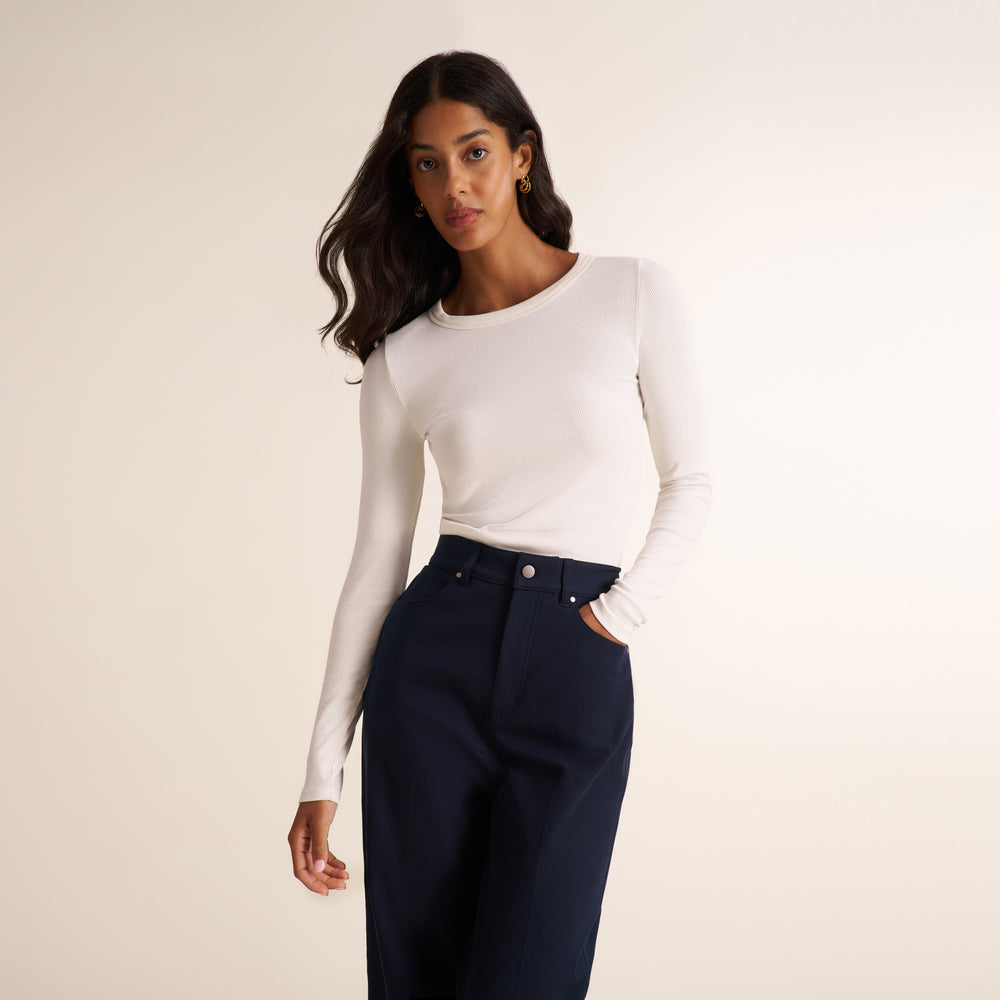 A woman modeling a white long-sleeve crewneck top and dark navy high-waisted trousers against a plain beige studio background.