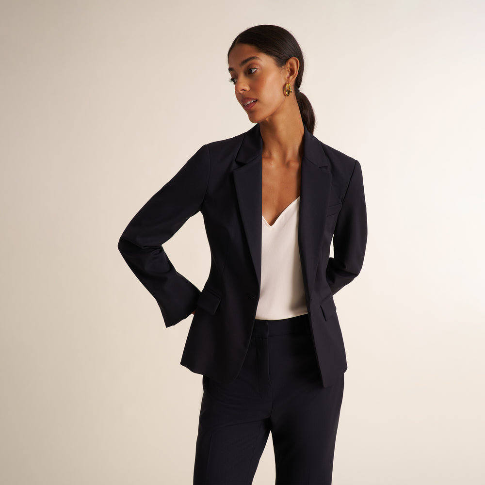 Woman wearing a black blazer, wide leg black suit pants, and a v-neck white tank top standing on a beige background
