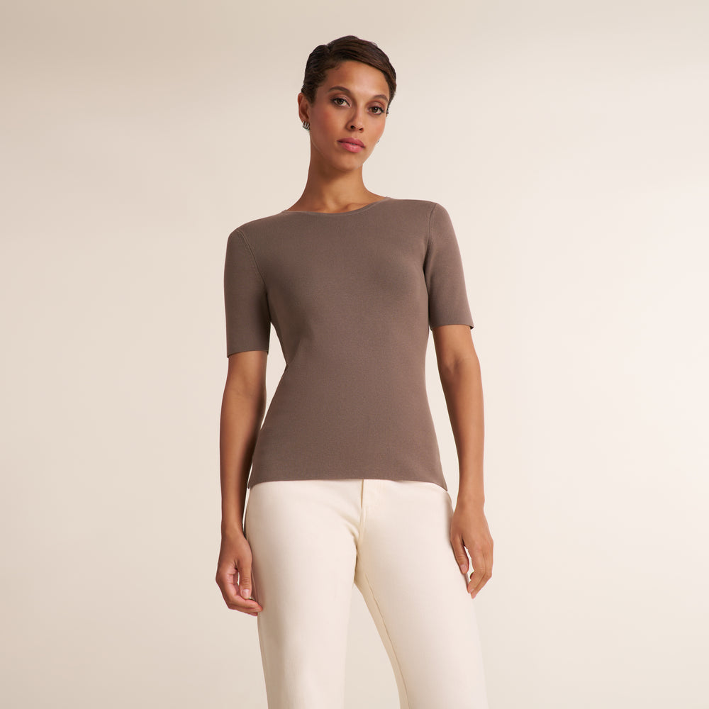 A woman modeling a short-sleeve ribbed knit top in taupe brown with off-white pants, standing against a plain light beige studio background.