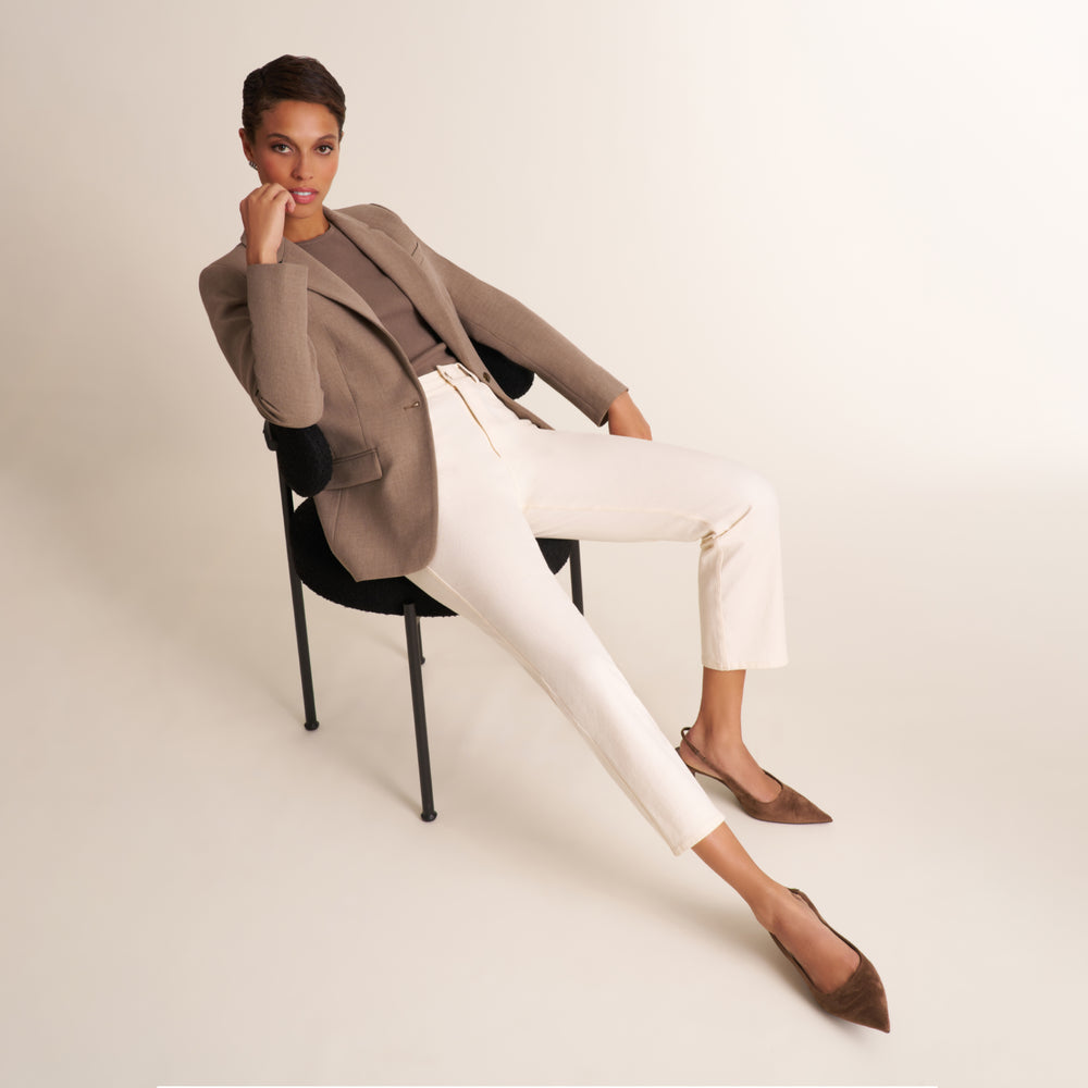 A model sits on a black chair against a warm off-white studio background, wearing a taupe blazer, a brown top, white cropped trousers, and brown pointed-toe flats.