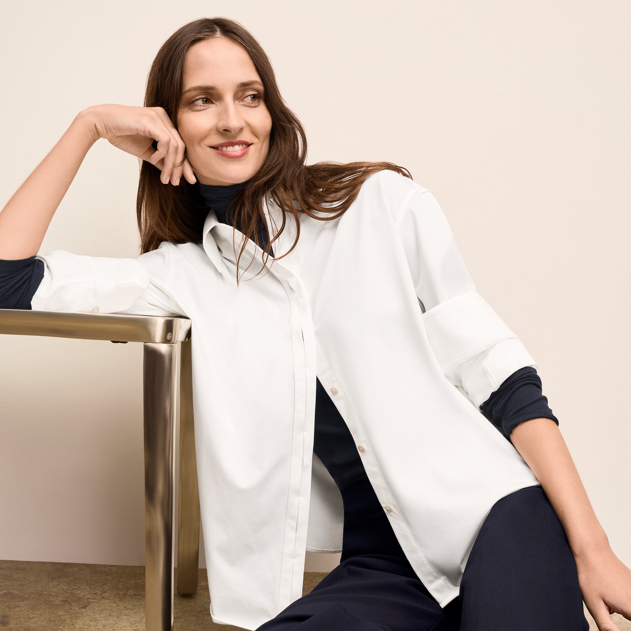 Woman in a white shirt and dark pants sitting on a chair against a plain background