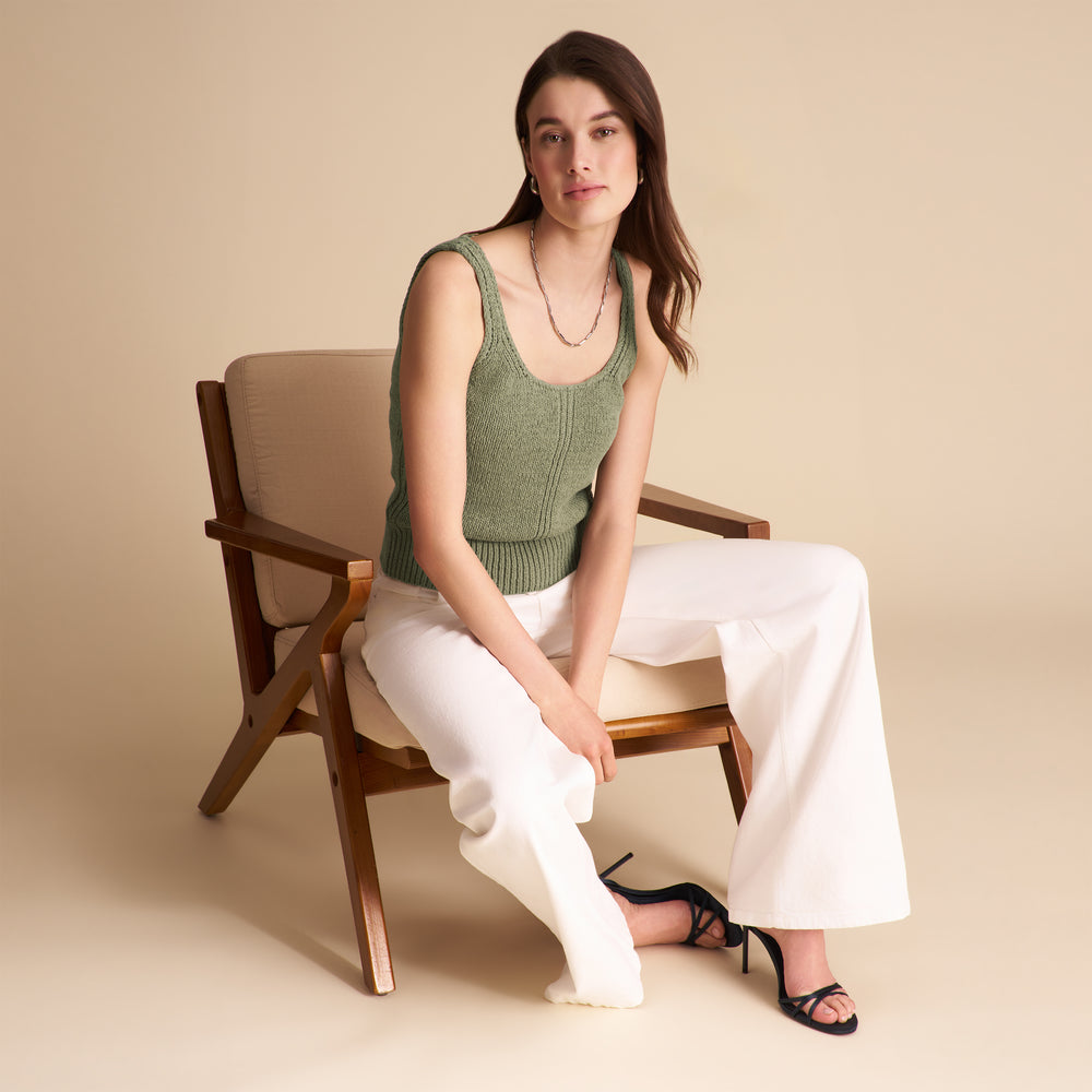 Woman sitting on a wooden chair wearing a green sleeveless top and white pants against a beige background