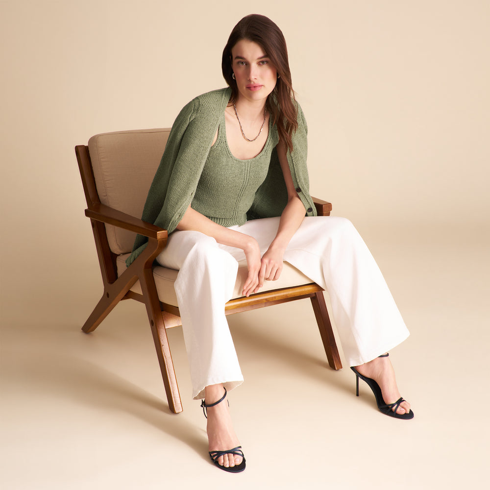 Woman sitting on a wooden chair wearing a green knit top and white pants against a beige background