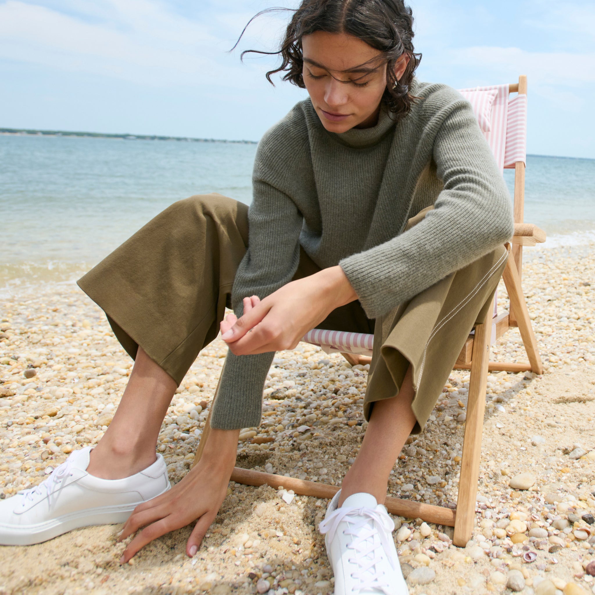 mood image of a woman sitting on a beach wearing the arbus sweater in dark moss