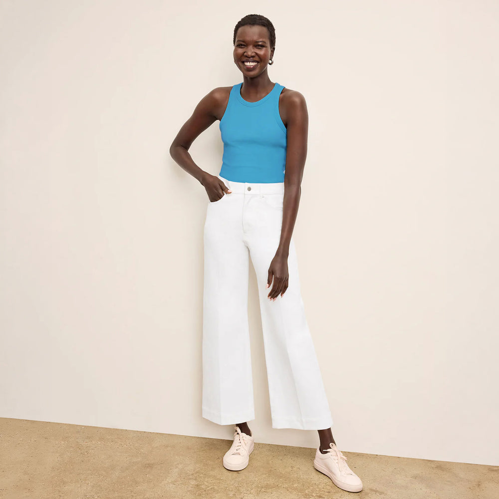 Woman wearing a blue tank top and white pants against a beige wall.