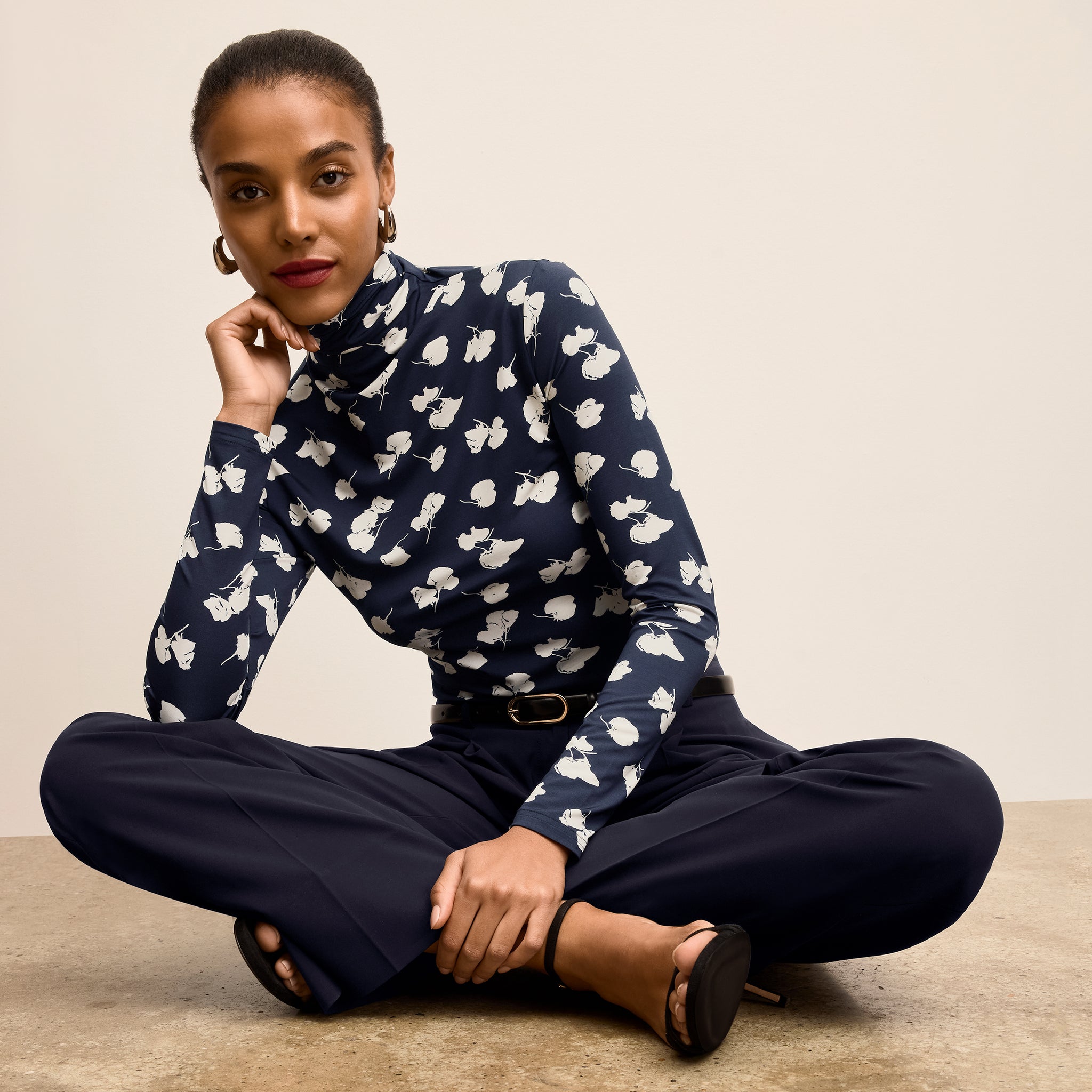 Woman wearing a navy blue turtleneck with white floral pattern and dark pants sitting on a beige floor 