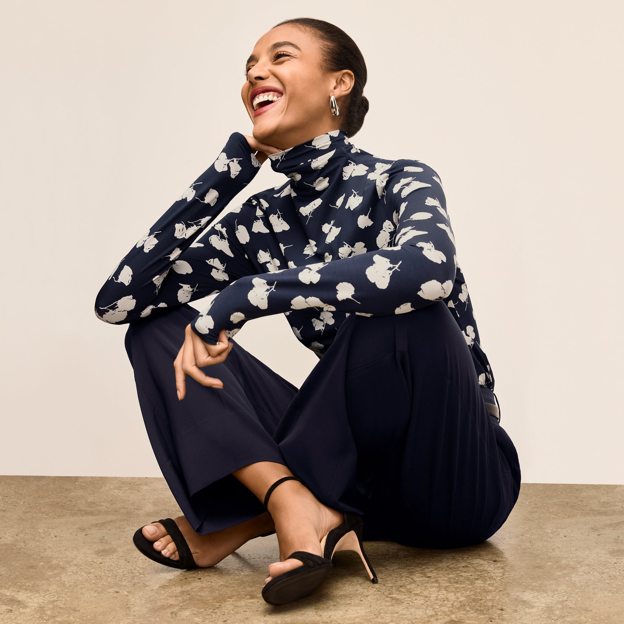 Woman wearing a navy floral blouse and dark blue skirt sitting on the floor against a beige wall.