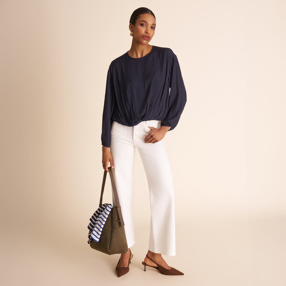 Woman wearing a navy top and white pants holding a green bag on a beige background
