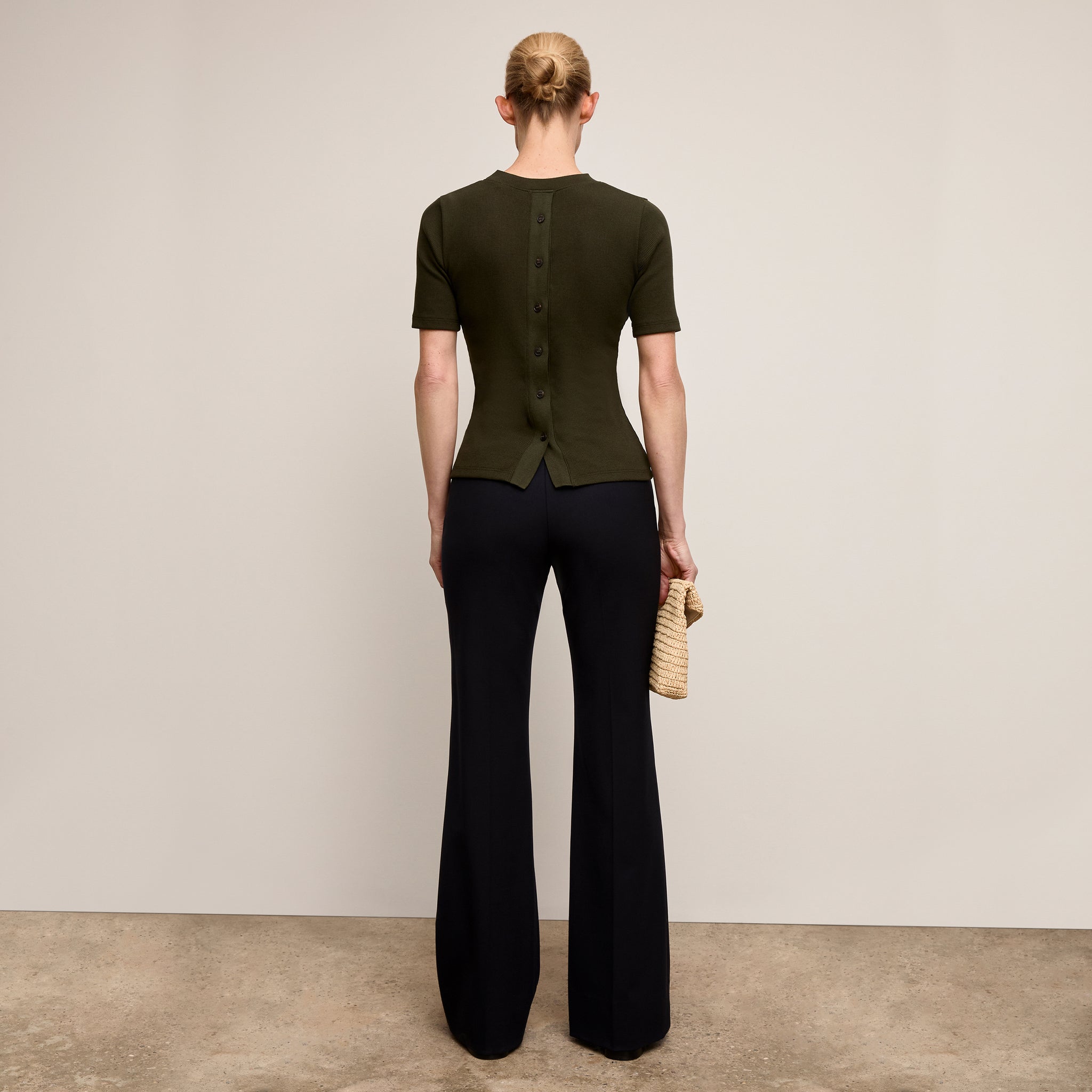 Person wearing a green top and black pants holding a straw bag against a plain background