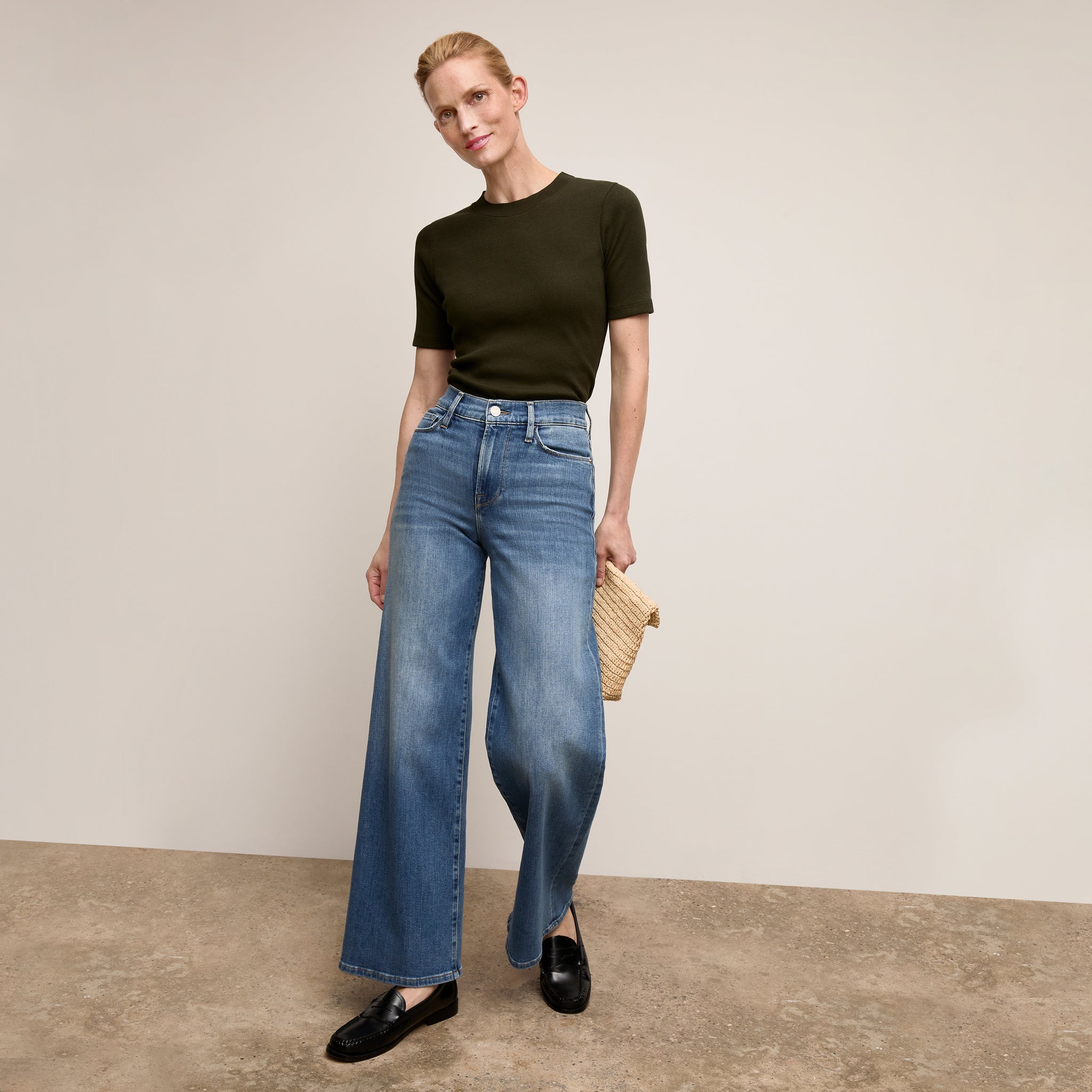 Woman wearing a black top and blue jeans holding a straw bag against a plain background
