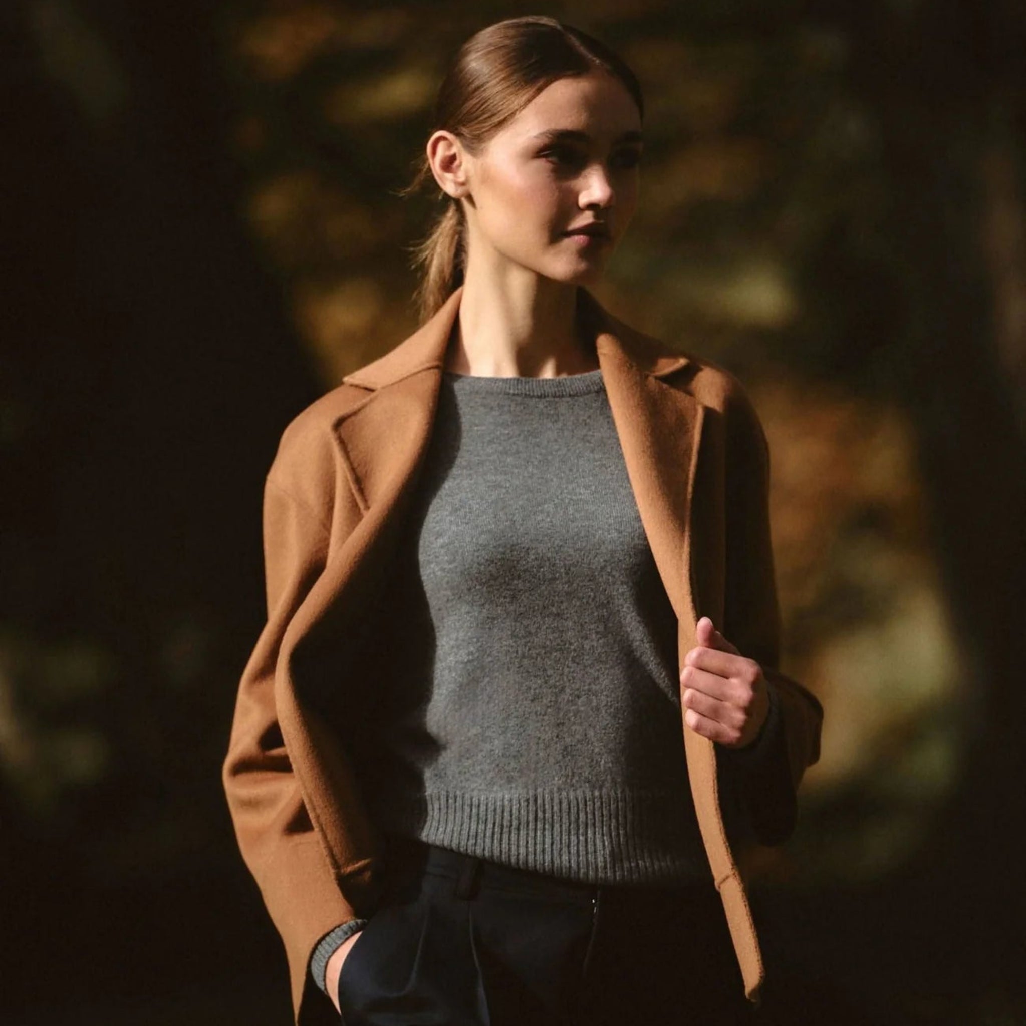 Woman wearing a brown coat over a gray sweater in a forest setting