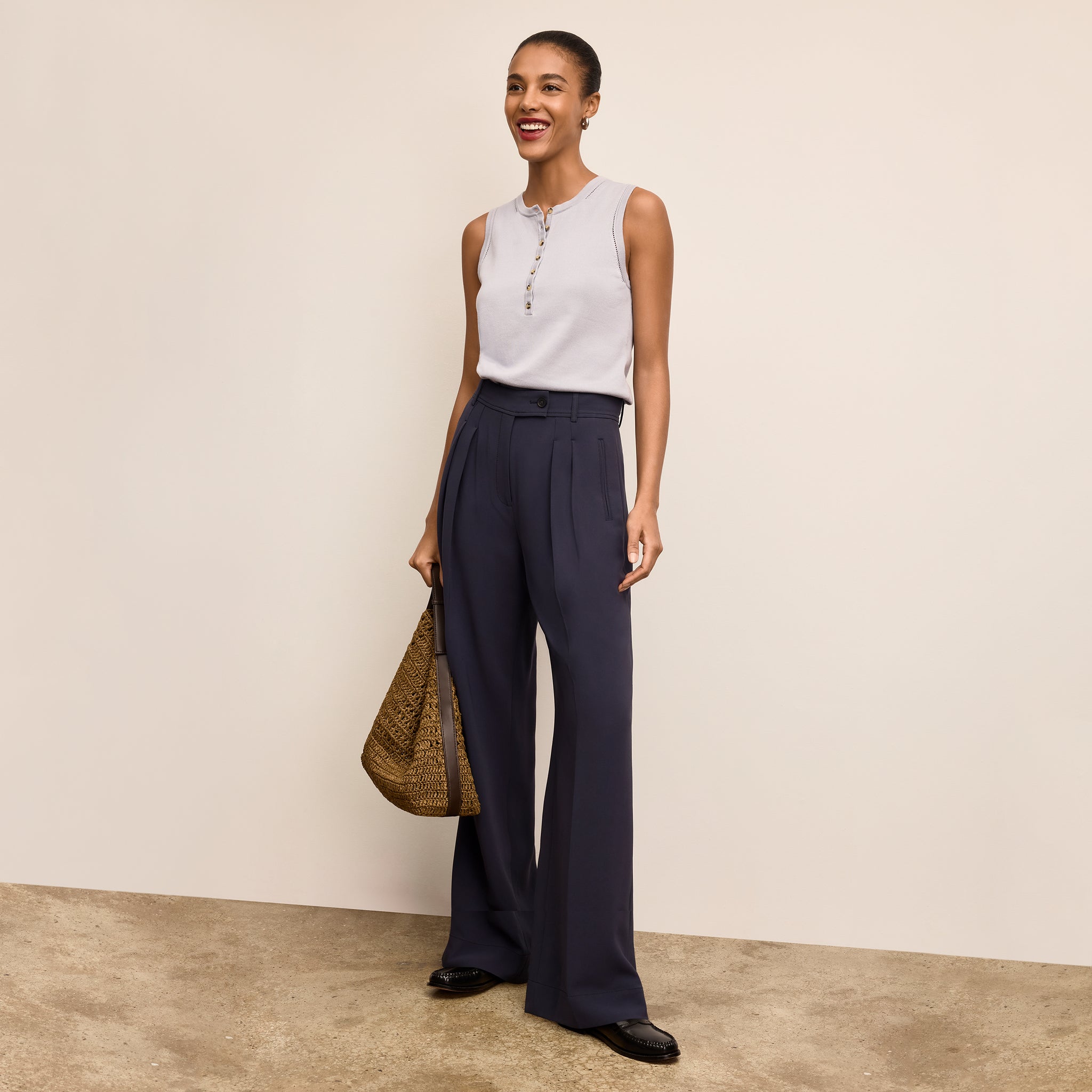 Woman wearing a sleeveless top and wide-leg pants, holding a bag against a plain background