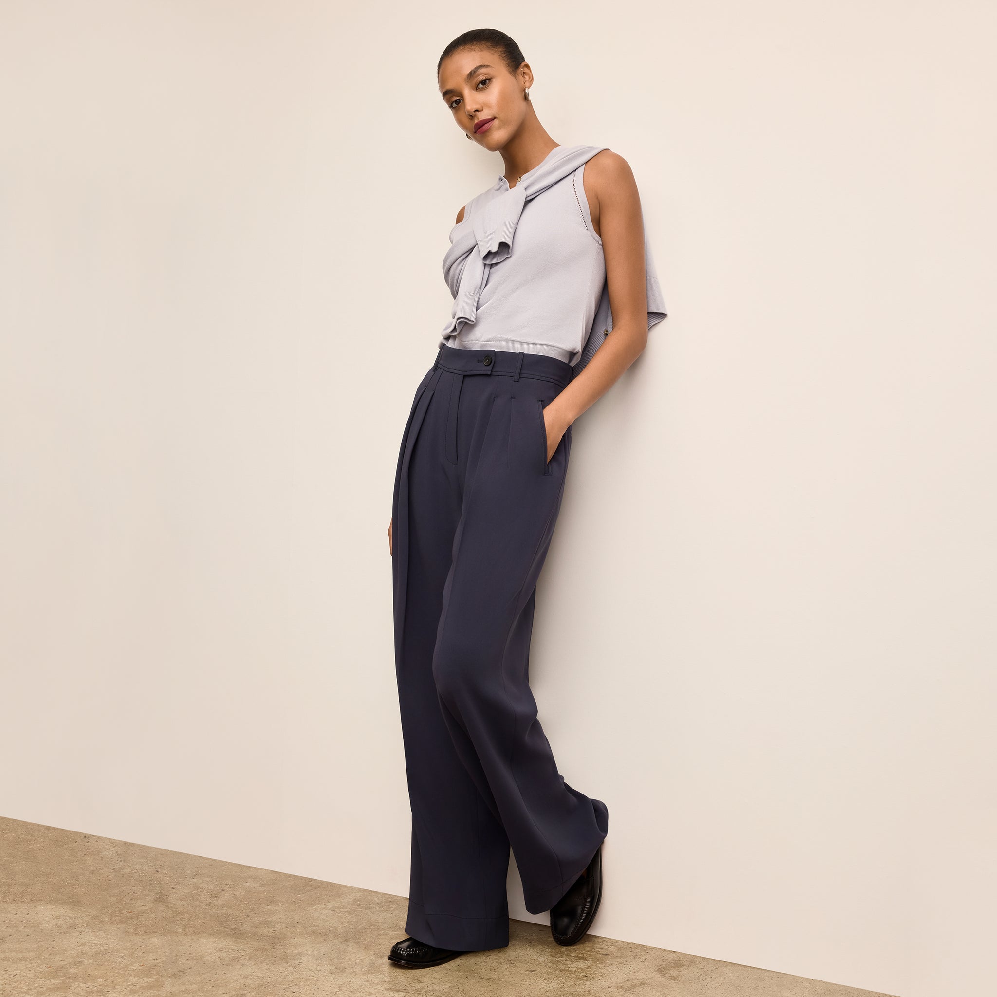 Woman wearing a light gray sleeveless top and dark blue high-waisted pants against a beige wall.