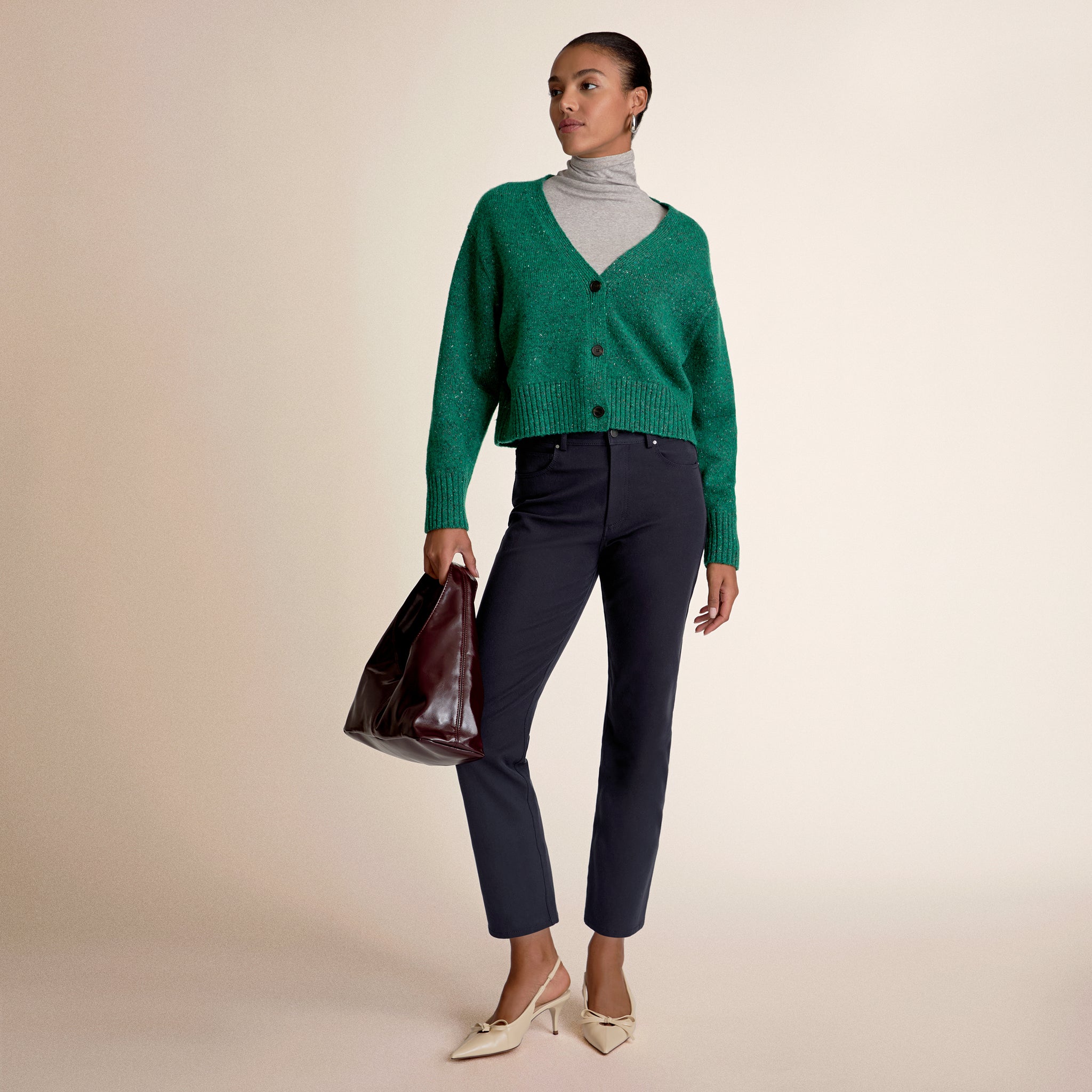 Woman wearing a green cardigan, gray turtleneck, and dark pants holding a brown bag on a beige background