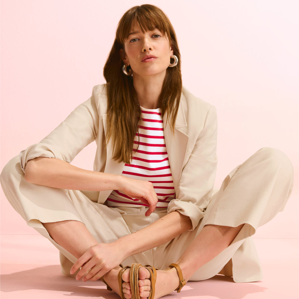 Woman sitting on a pink background wearing a beige blazer, striped shirt, and wide-leg pants.