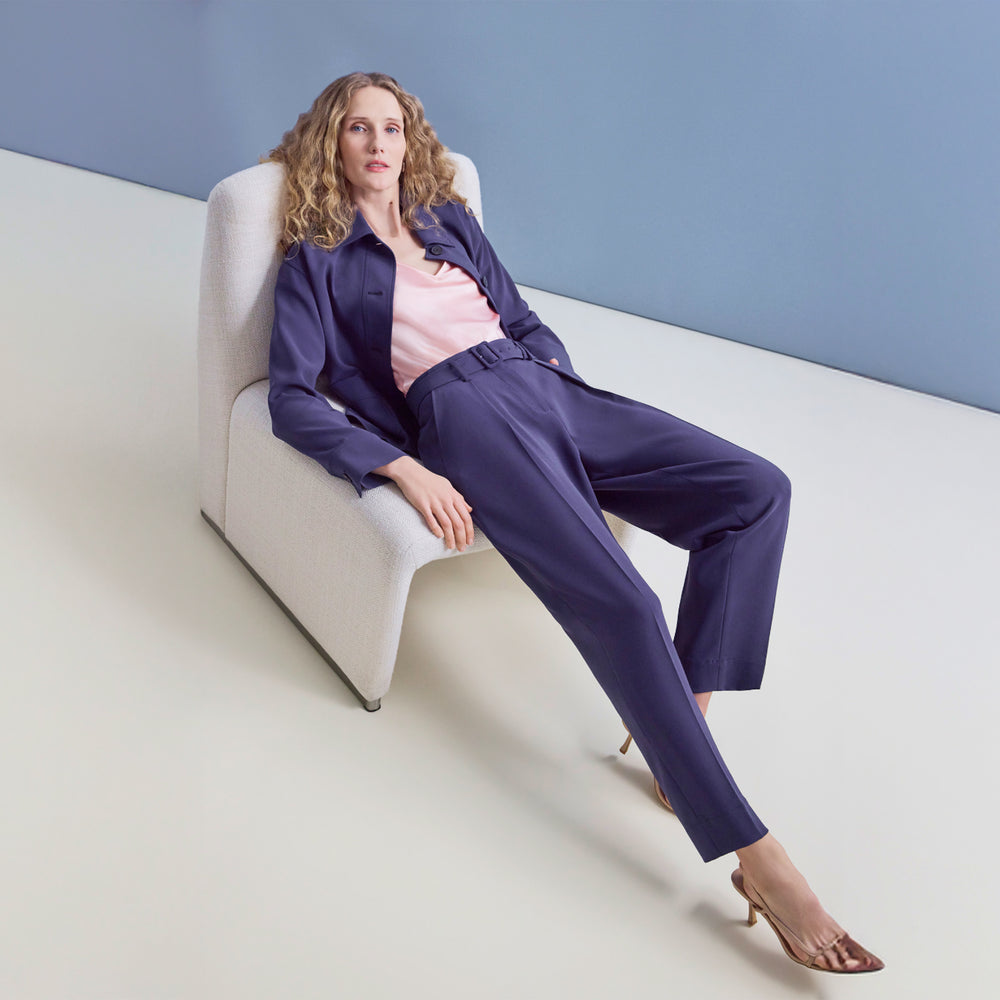 Woman in a navy suit sitting on a white chair against a light blue wall.