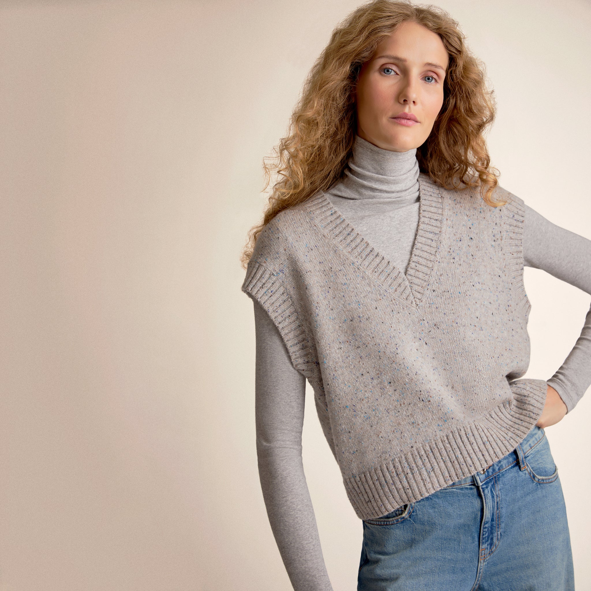 Person wearing a gray sweater vest and blue jeans on a beige background 