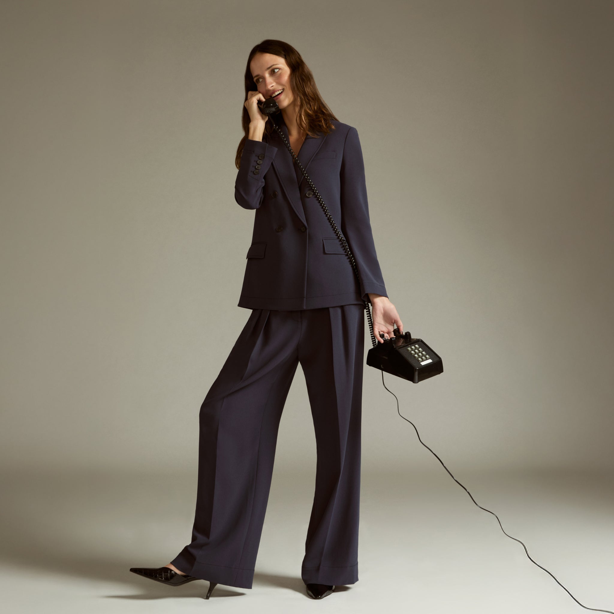 Woman in a navy suit holding a vintage phone with a long cord against a plain background