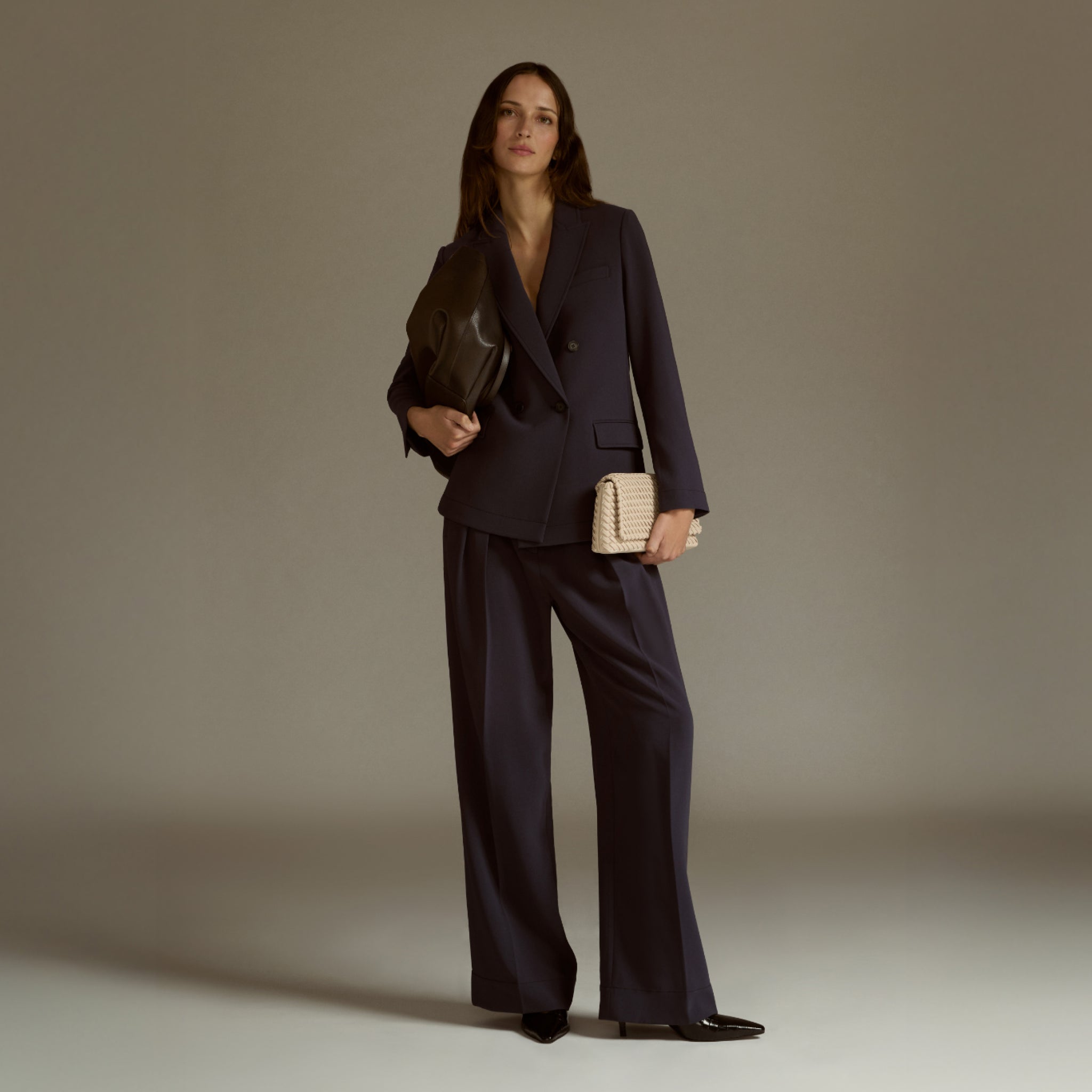 Woman in a dark suit holding a beige handbag against a neutral background 