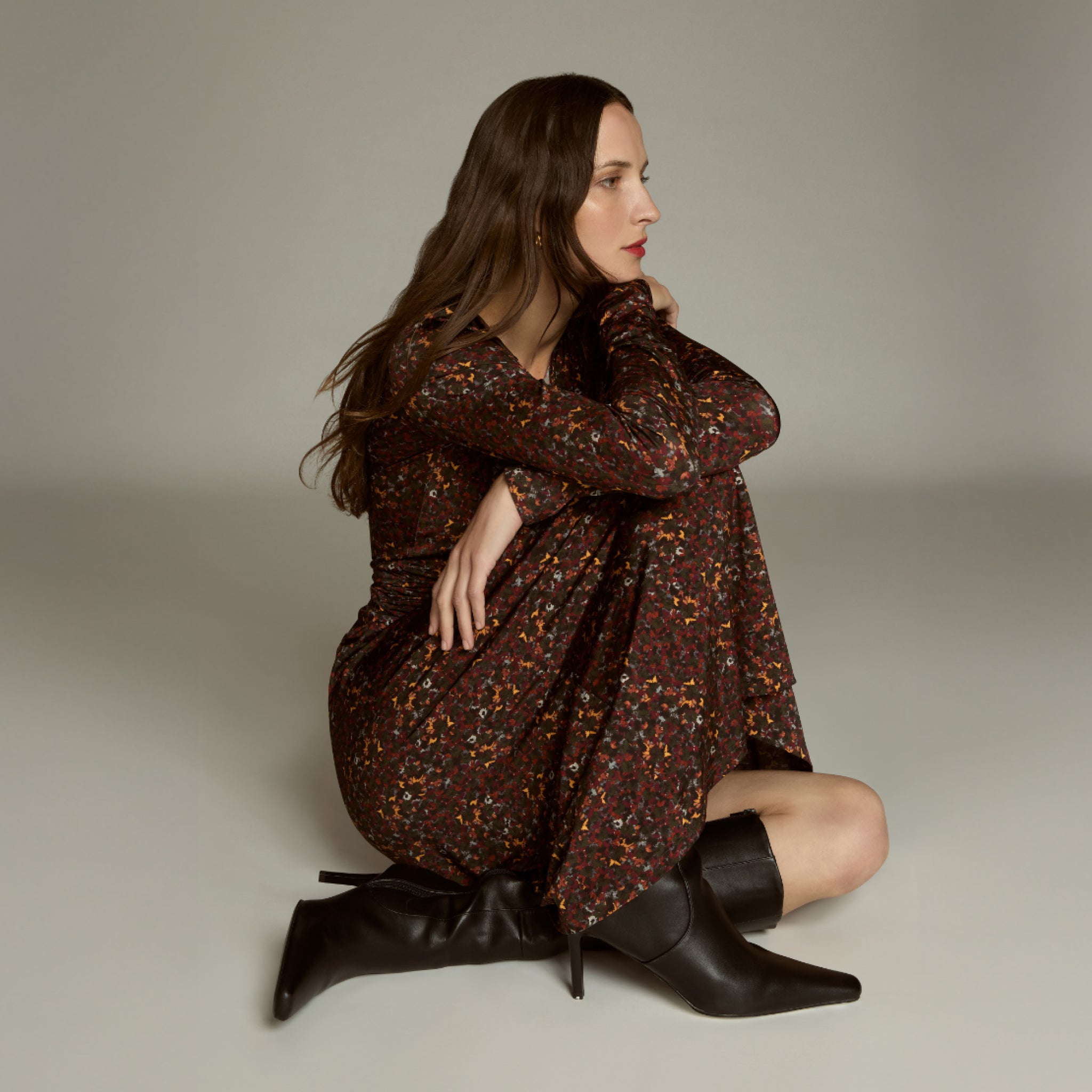 Woman in a patterned dress and black boots sitting on a neutral background