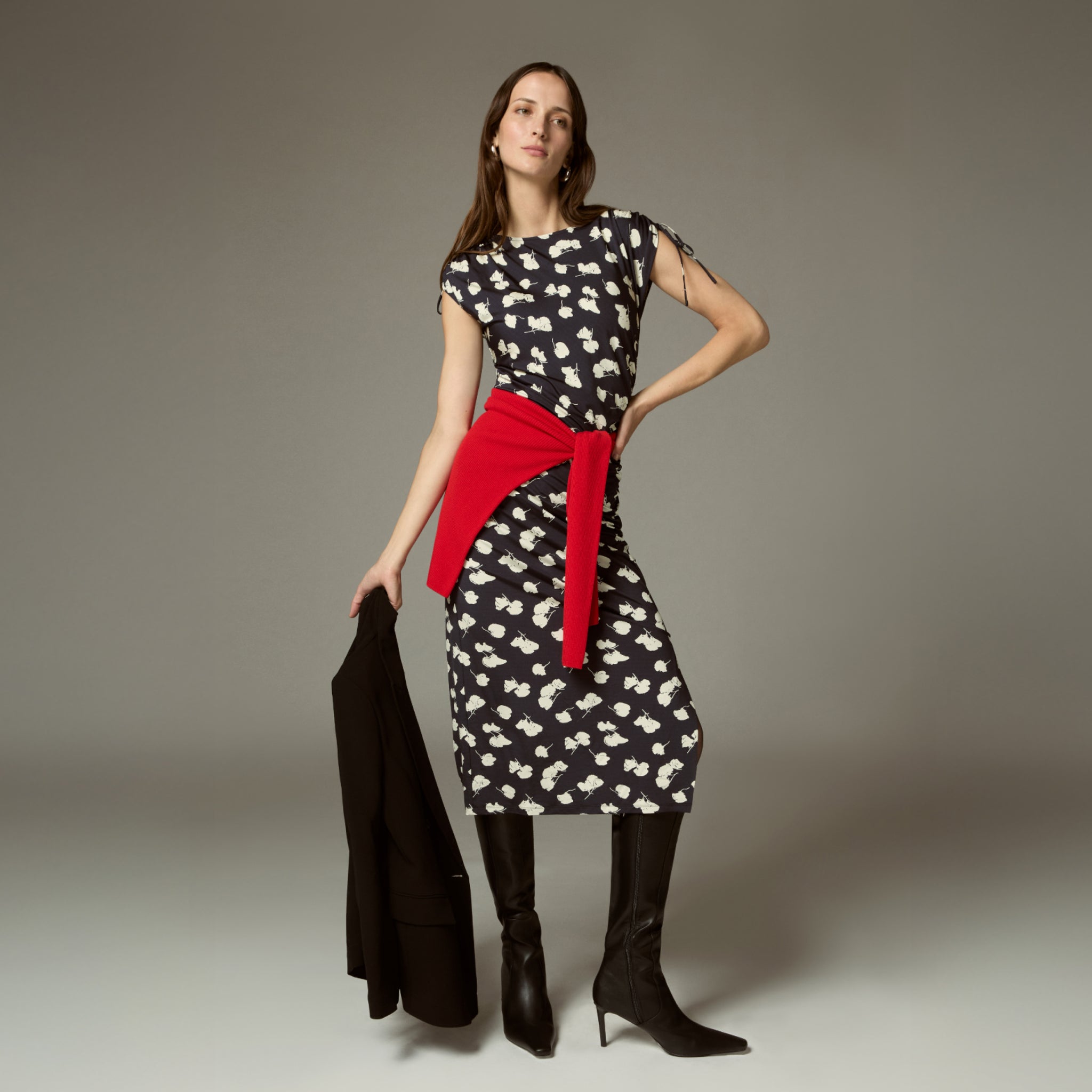 Woman wearing a black dress with white floral pattern and red belt, holding a black jacket against a neutral background.