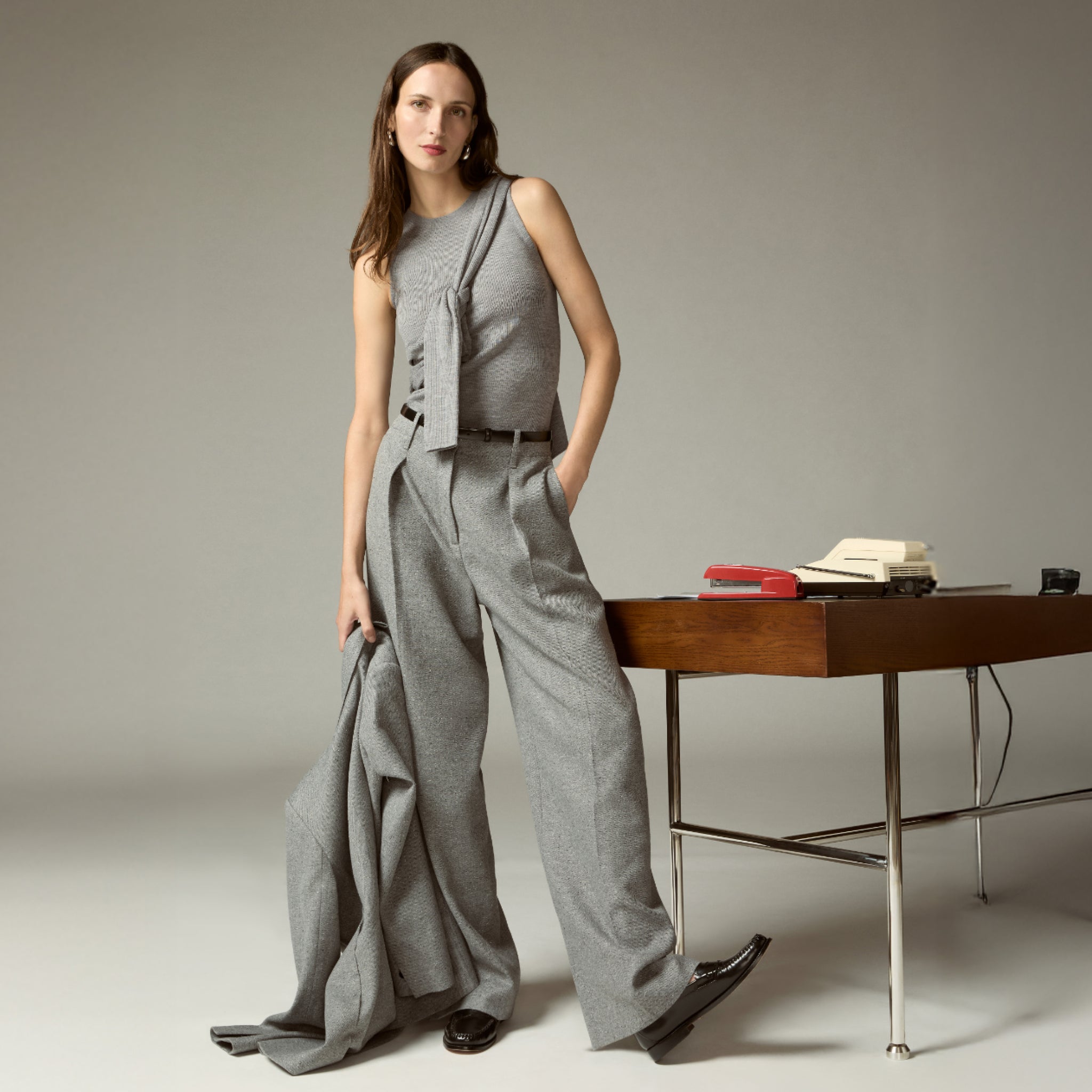 Woman in gray outfit standing next to a wooden desk with books and a phone on it.
