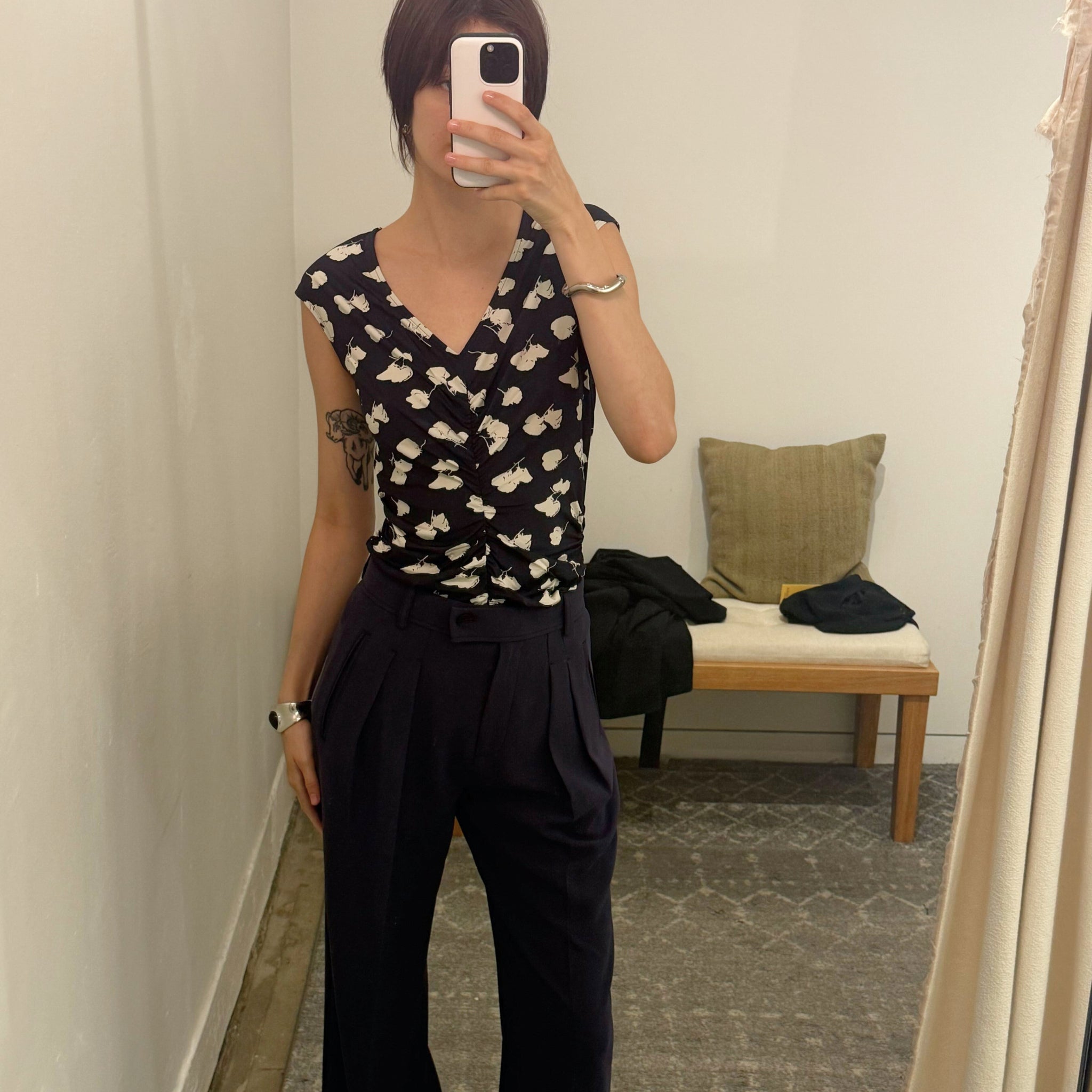 Person wearing a black and white patterned top and dark pants in a room with a bench and pillow.