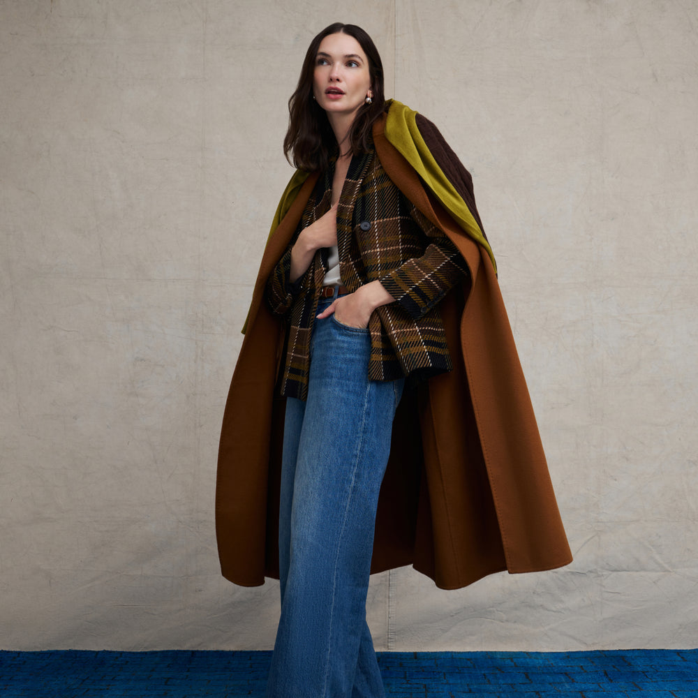 Woman wearing the O'Hara Blazer in Heritage Plaid with a brown suede long coat over it and wide leg jeans on a beige background with blue floor