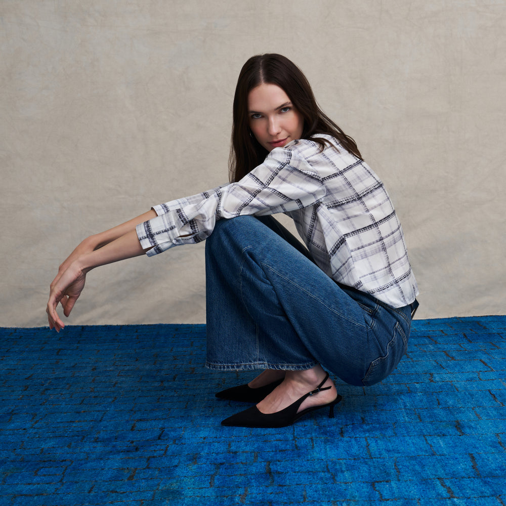 Woman wearing a white and gray plaid silk long sleeve boat neck top and wide leg jeans on a blue carpet floor and against a beige background