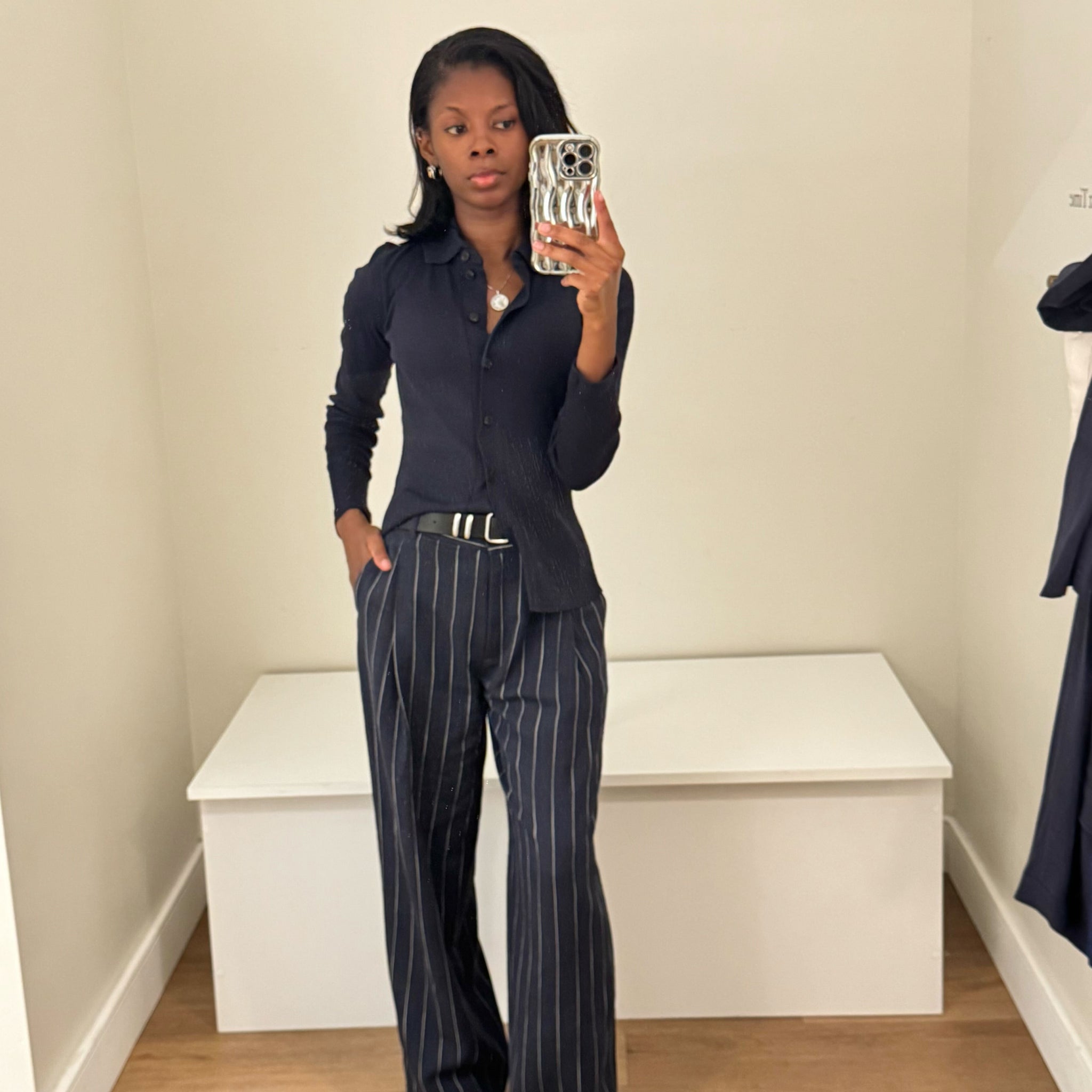 Person taking a mirror selfie wearing a dark blue shirt and pinstripe pants in a minimal room.