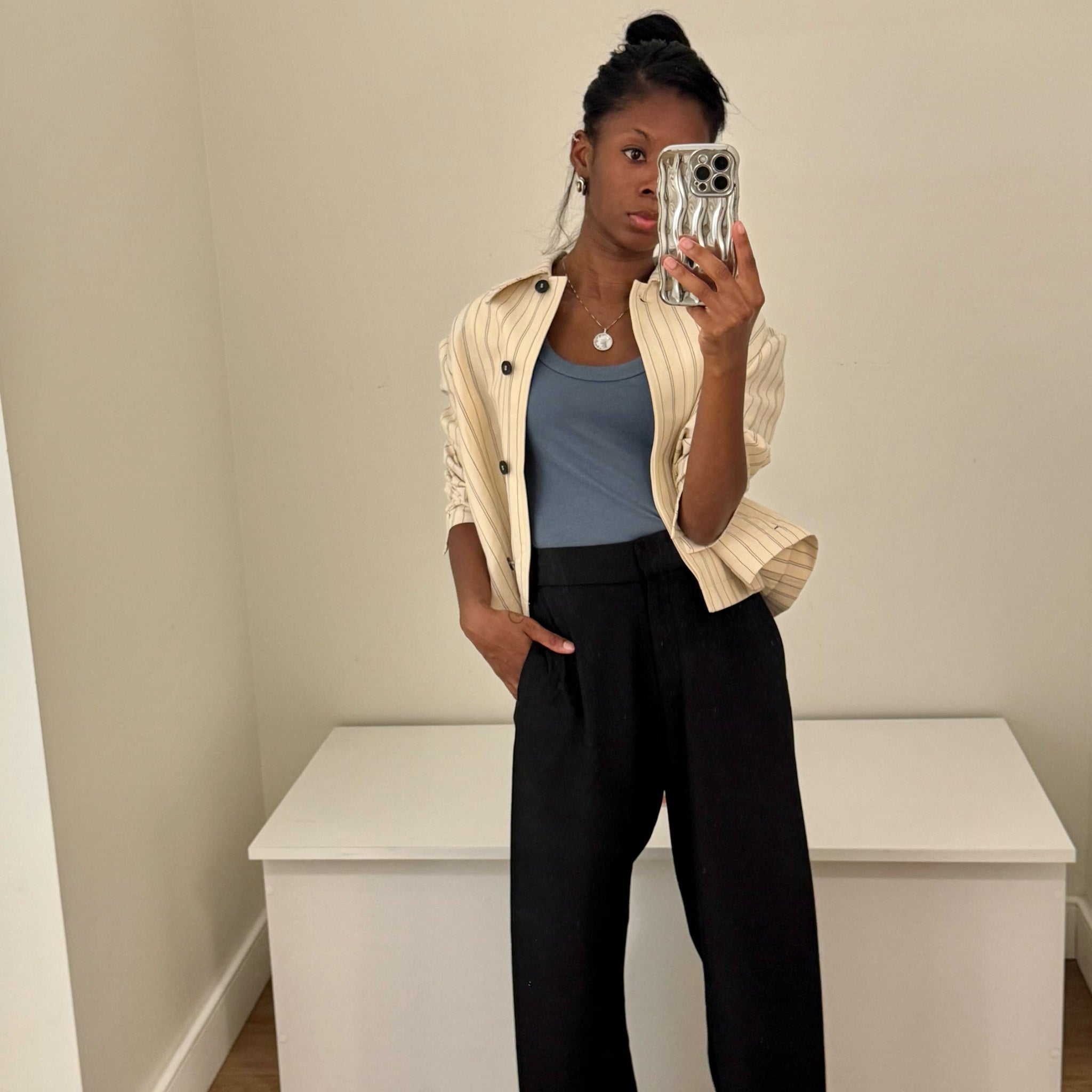 Person taking a mirror selfie wearing a beige jacket, blue top, and black pants in a minimalistic room.