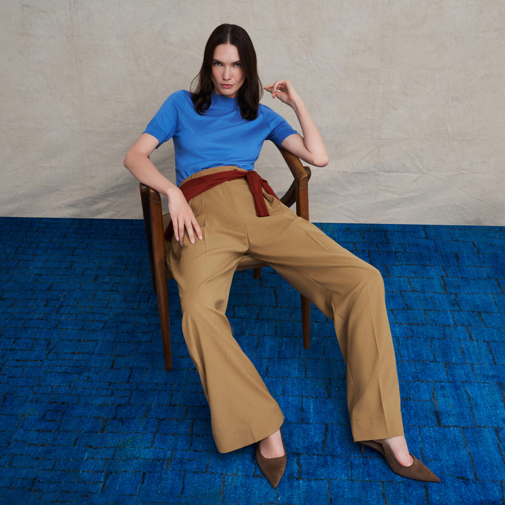 Woman wearing a short sleeve blue splendor sweater and beige wide leg pants on a blue carpet floor and against a beige background
