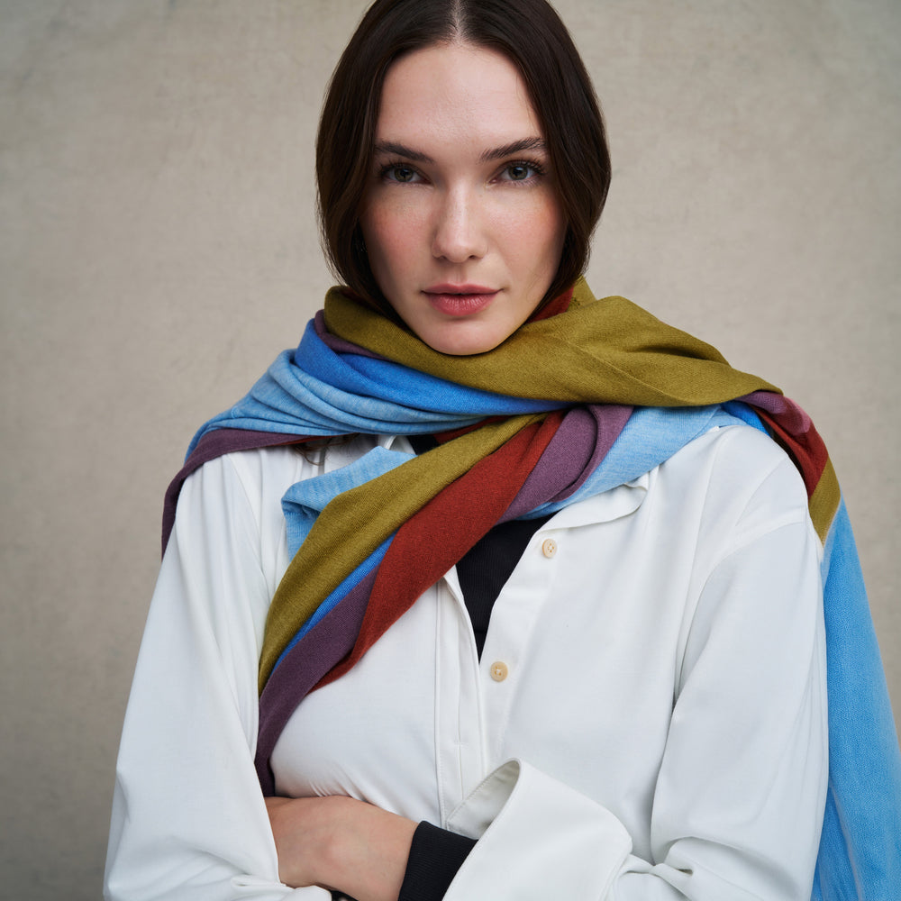 Woman wearing a white long sleeve button down and skinny black pants wth multiple sweaters wrapped around her neck like a scarf on a blue carpet floor and against a beige background