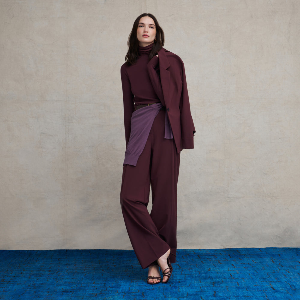 Woman wearing an aubergine turtleneck long sleeve ribbed top with a matching aubergine jacket and wide leg pant suit with a purple sweater tied around her waist while standing on a blue carpet floor with a beige background