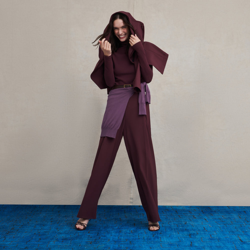 Woman wearing an aubergine turtleneck long sleeve ribbed top with a matching aubergine jacket and wide leg pant suit with a purple sweater tied around her waist while standing on a blue carpet floor with a beige background