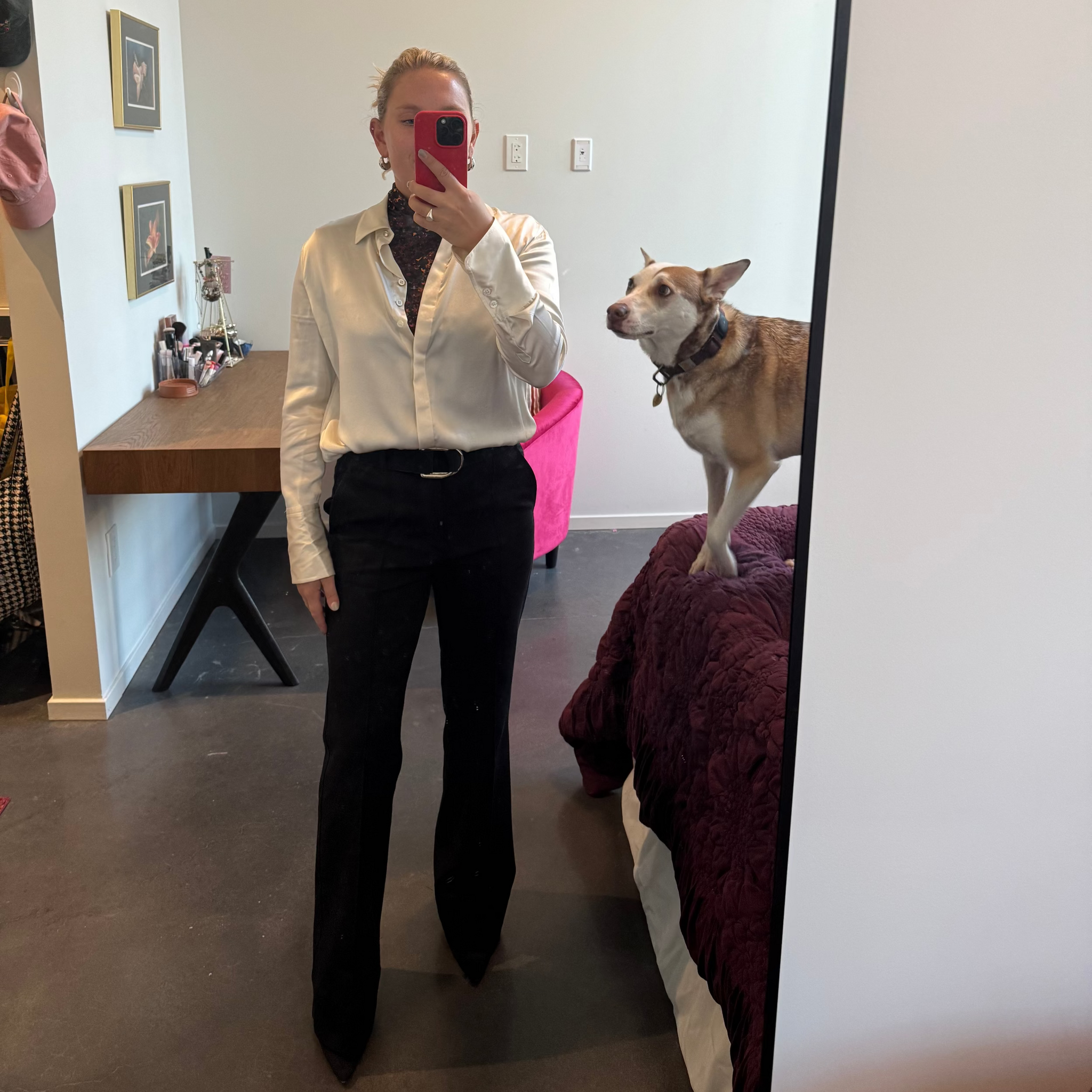 Person taking a mirror selfie wearing a white shirt and black pants, with a dog on a bed in the background.
