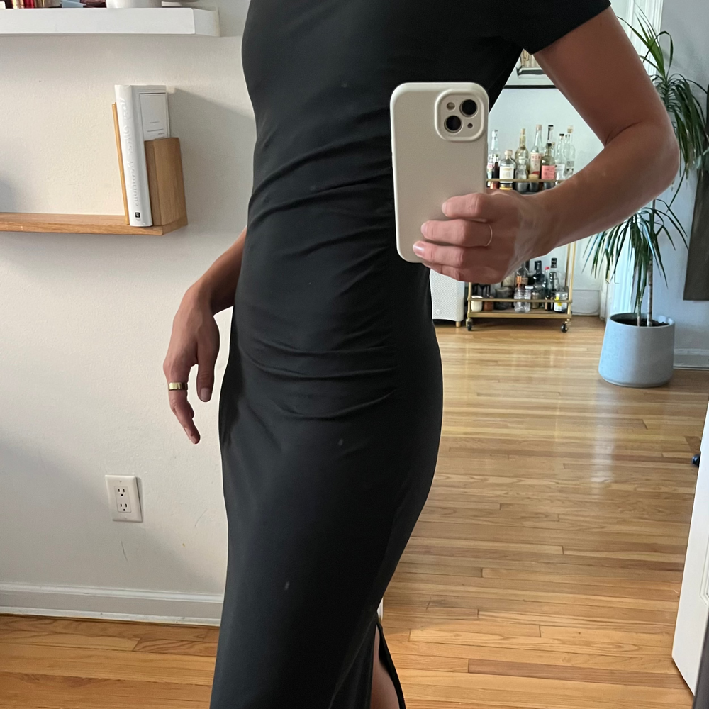 Person wearing a black dress with a slit, standing in a room with wooden flooring and shelves.