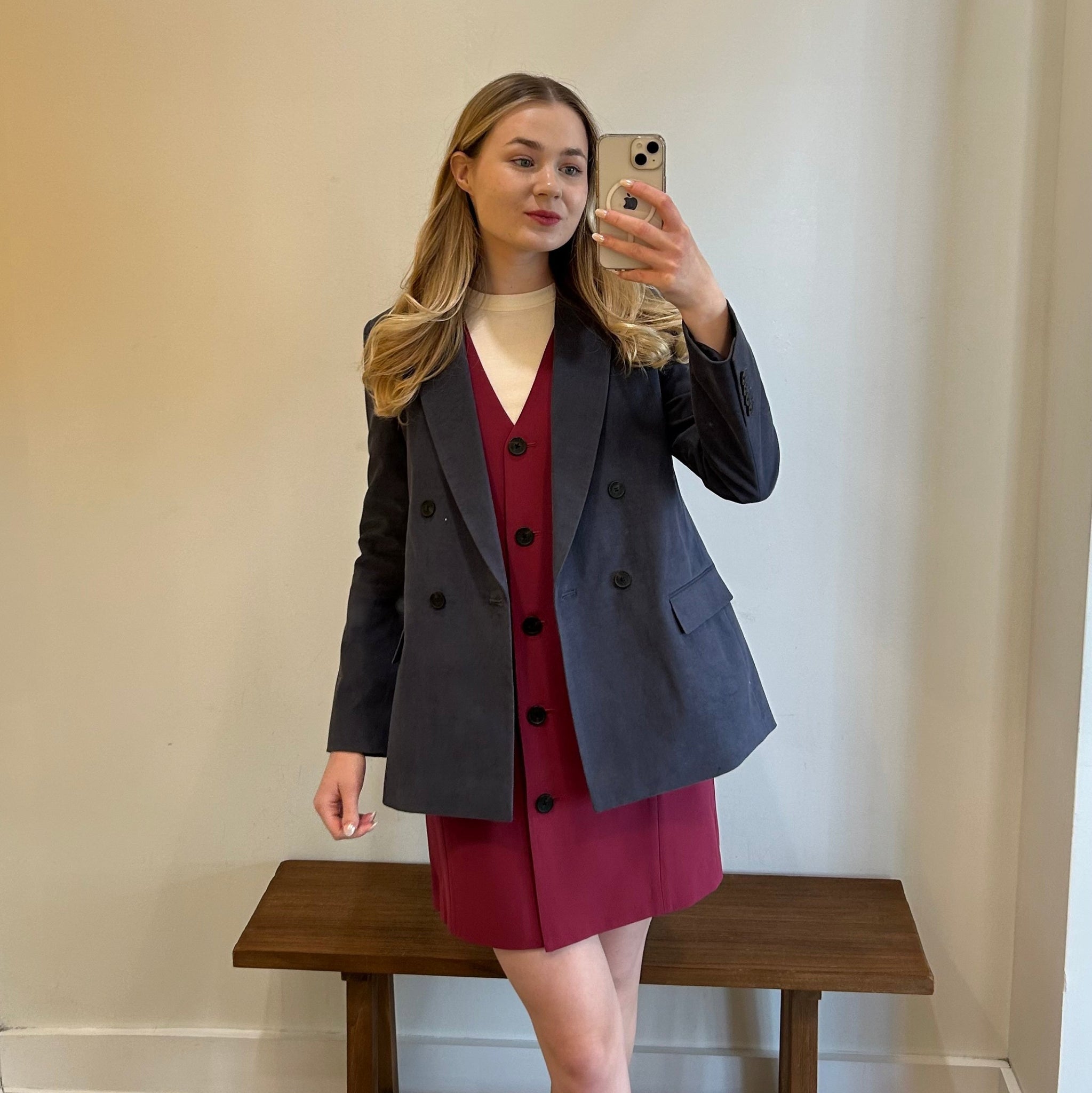 Woman taking a mirror selfie wearing a dark gray blazer over a red dress on a wooden bench.