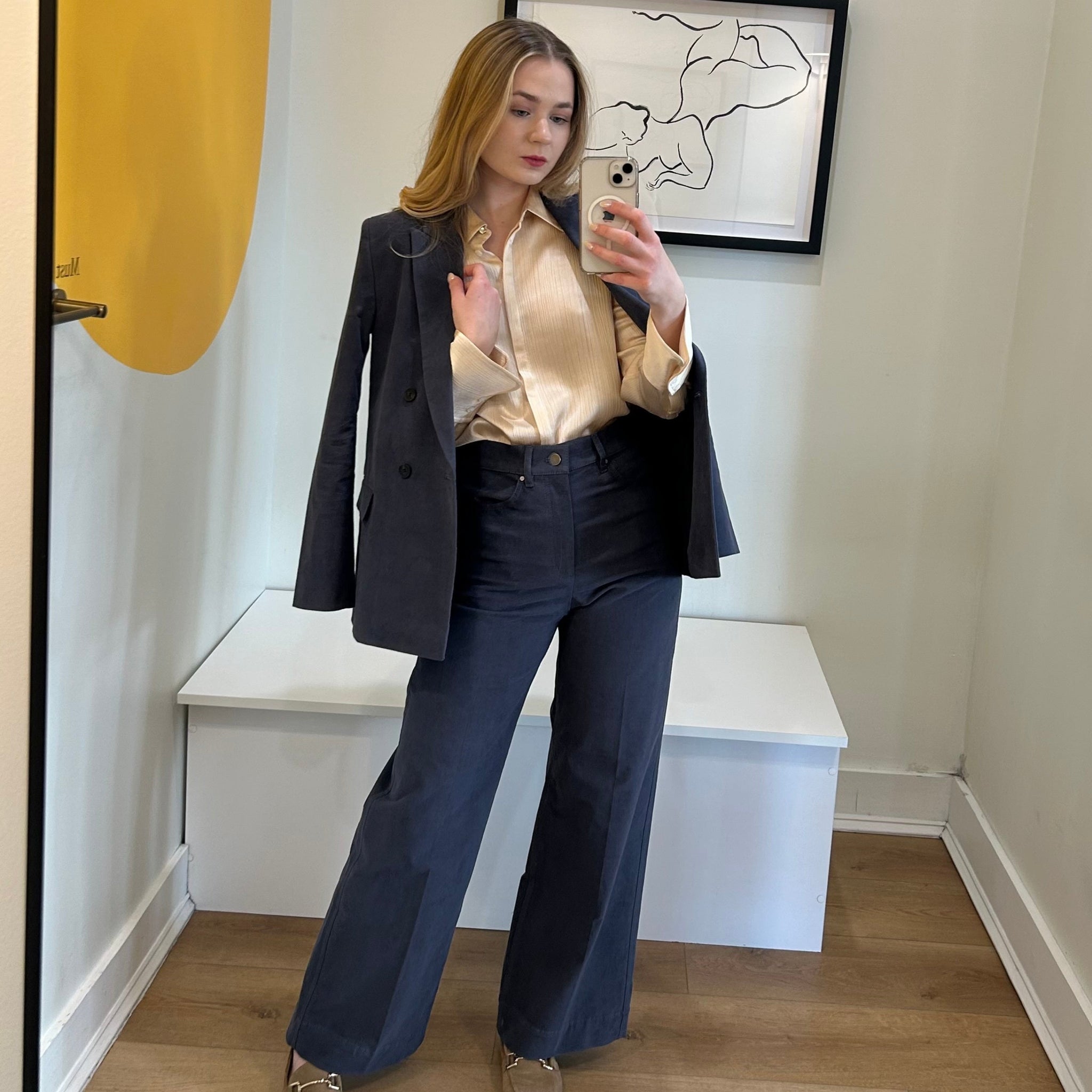 Woman taking a mirror selfie wearing a dark blazer, light blouse, and blue pants in a room with a yellow wall and white bench.