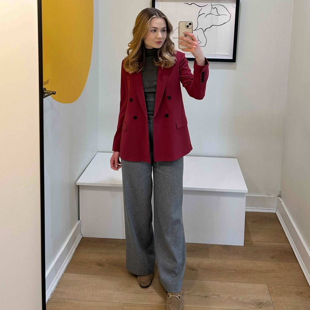 Person wearing a red blazer and gray pants taking a mirror selfie in a room with a yellow surfboard and abstract art on the wall.