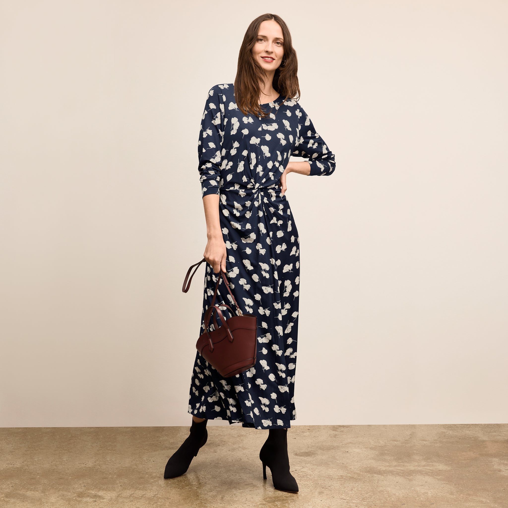 Woman wearing a navy dress with white floral pattern, holding a brown handbag against a beige background 