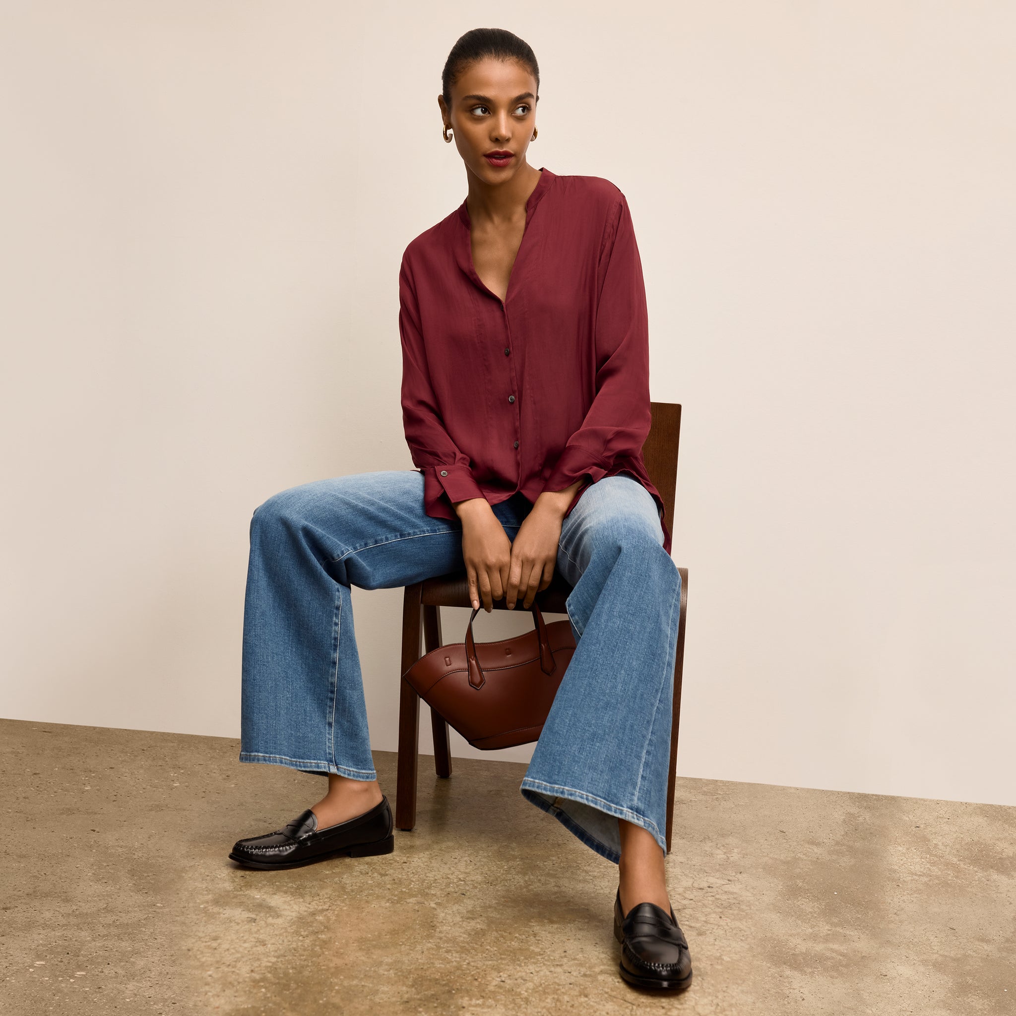Woman sitting on a chair holding a brown handbag, wearing a red blouse and blue jeans 